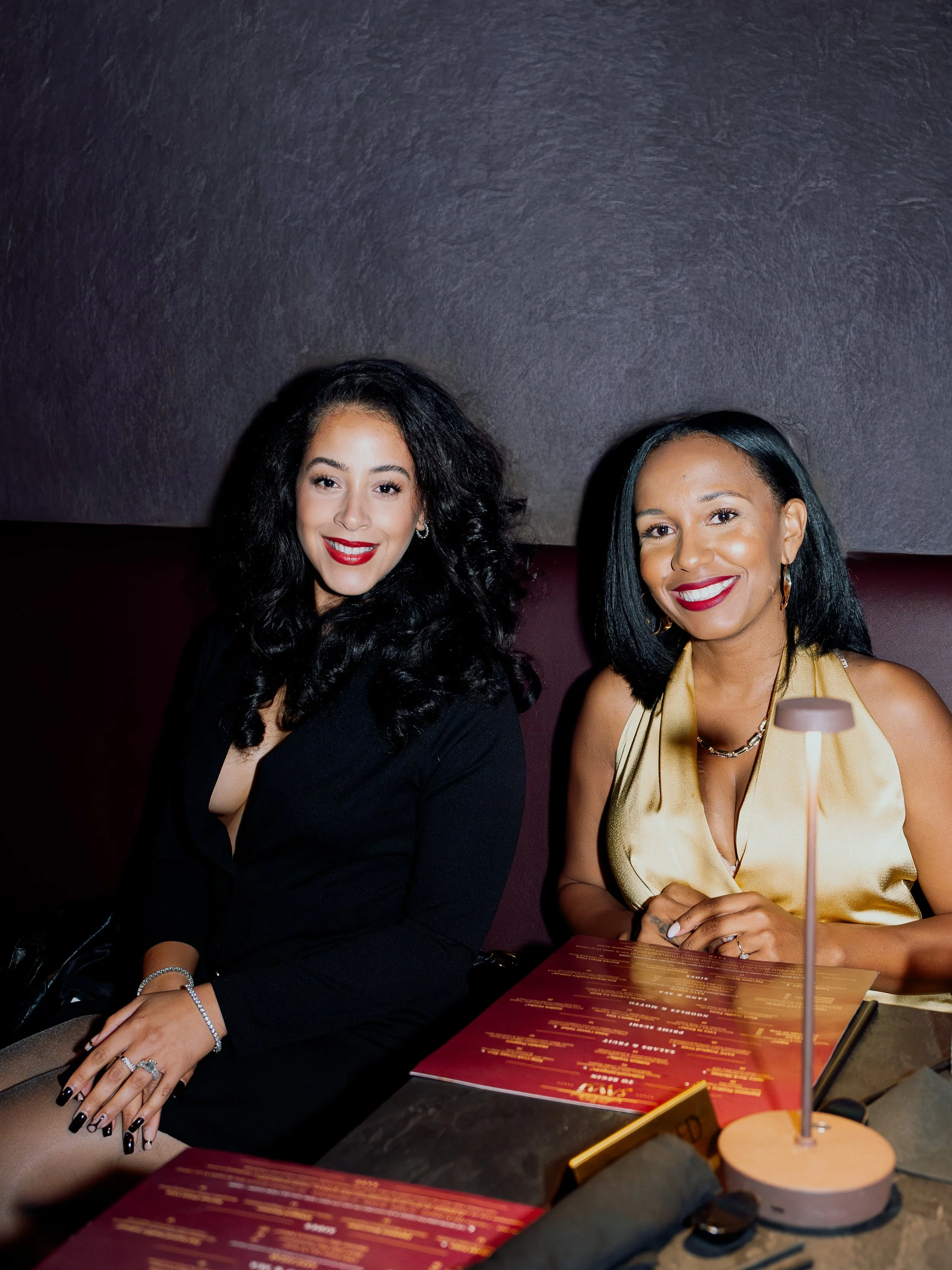 Two women smiling and sitting at a table in a dimly lit restaurant, with menus and a small lamp in front of them.