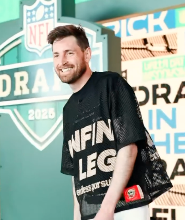 A man with a beard and short brown hair smiling at an NFL draft event in 2023, standing in front of a large NFL Draft sign, wearing a black mesh shirt with white text and a red wristband.