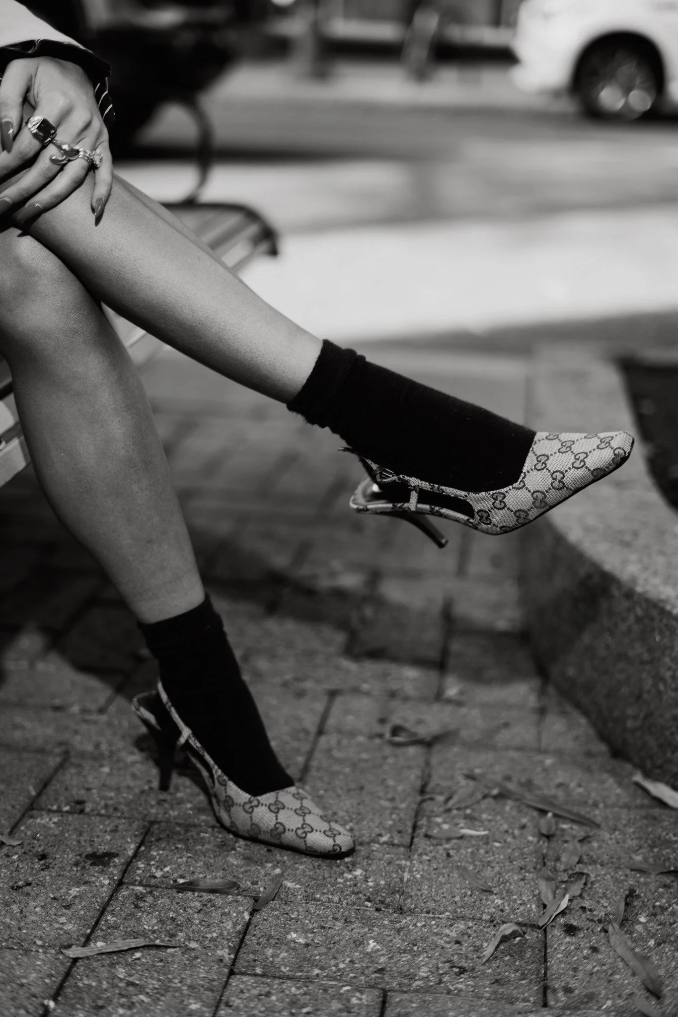 A person sitting on a bench with crossed legs, wearing patterned high heels, black socks, and rings on fingers, with a street scene in black and white in the background.