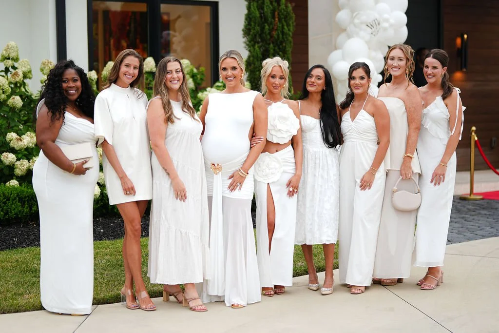 A group of ten women dressed in white outfits standing together outside in front of a building decorated with white flowers and balloons.