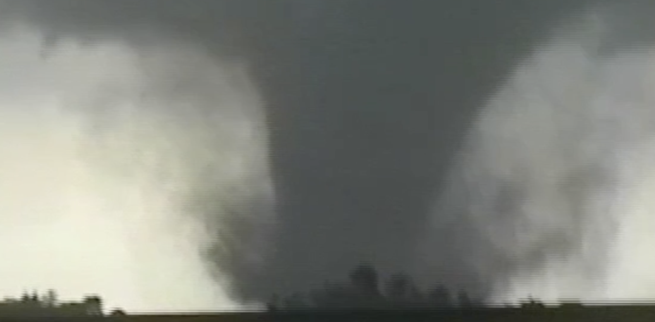 A large tornado touching down near a horizon of low trees and buildings, with dark storm clouds overhead.