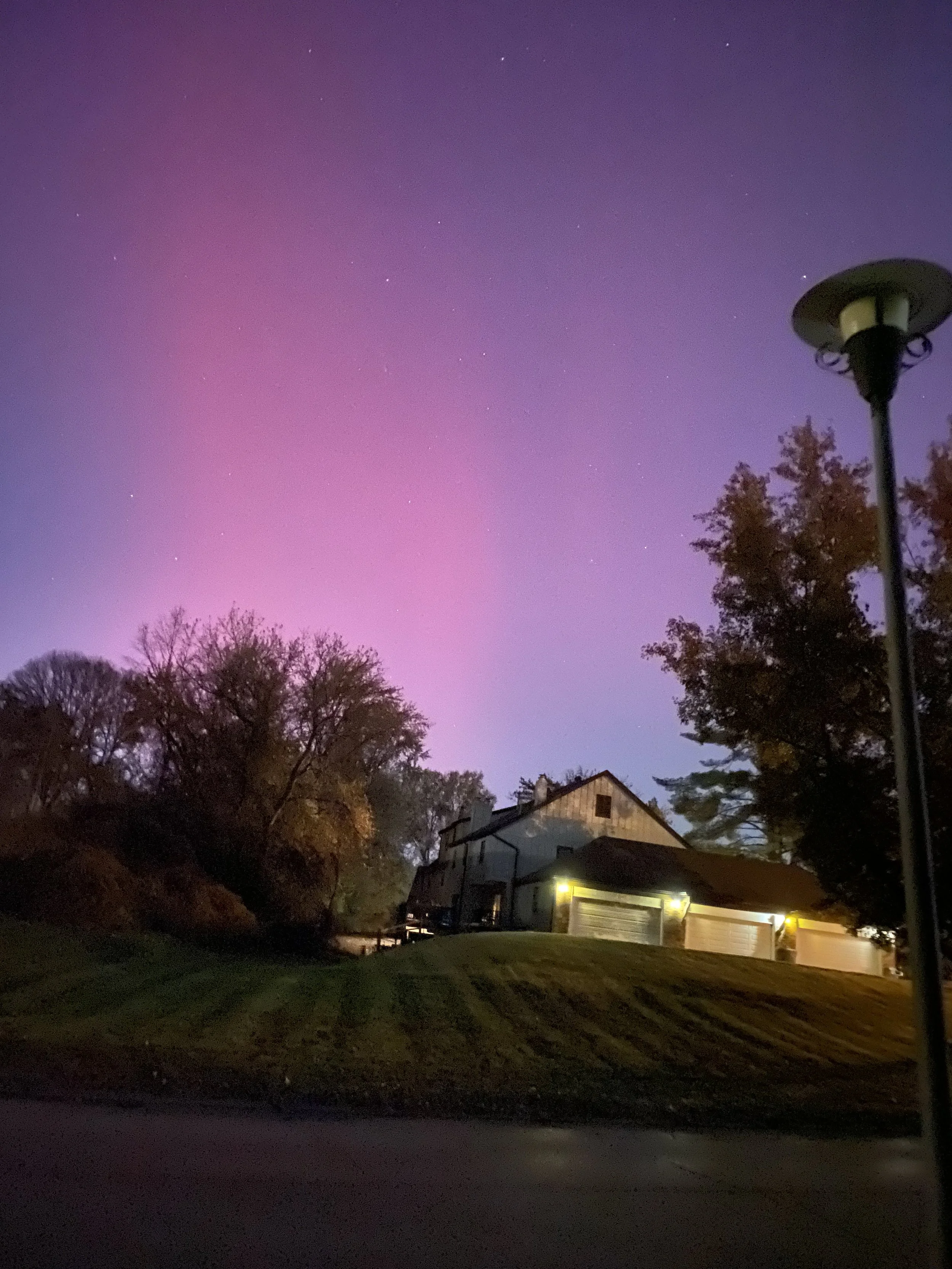Nighttime scene of a house with illuminated garage doors, trees, and a lamp post under a colorful, purple-pink gradient sky with visible stars.