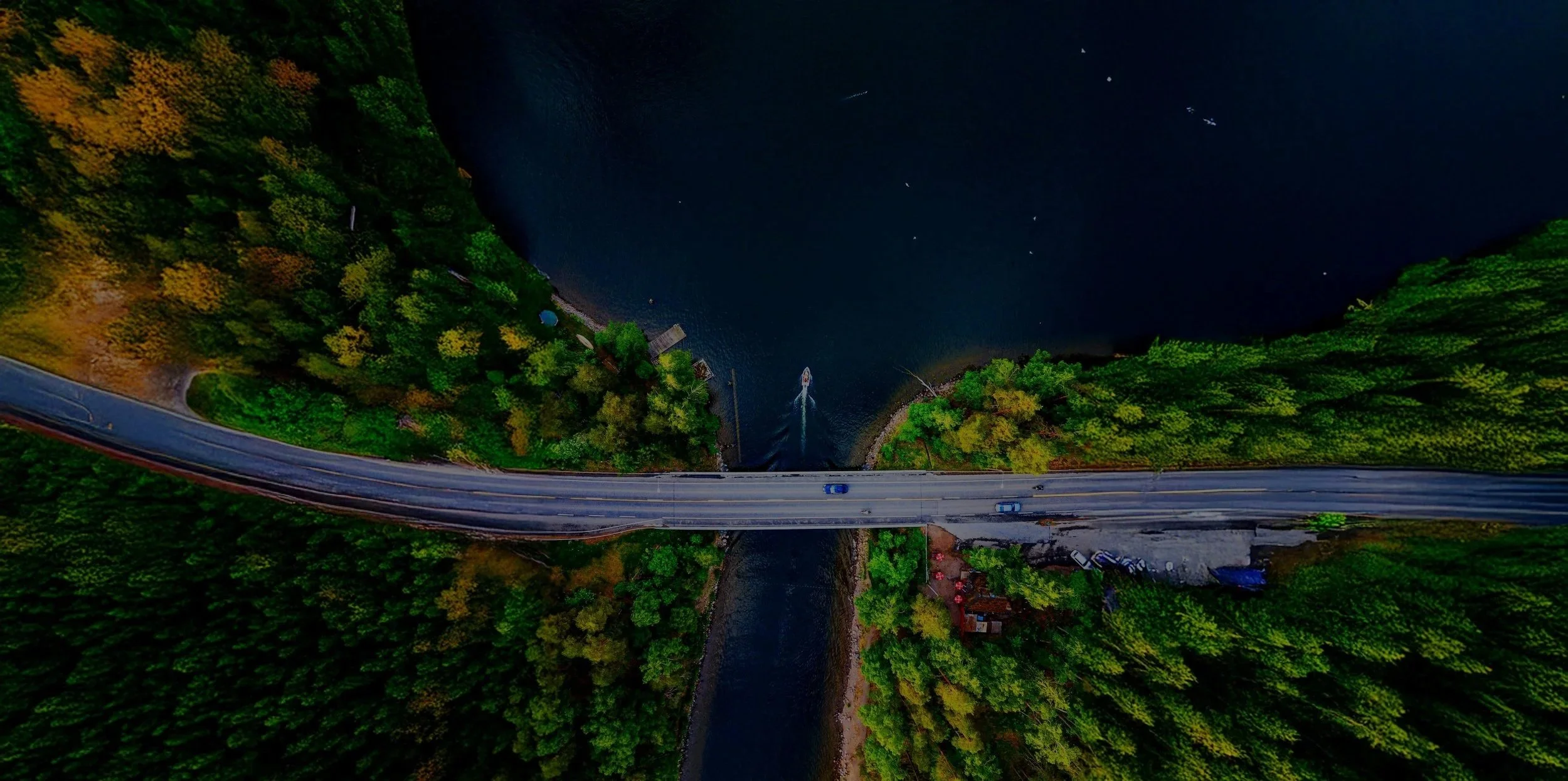 Aerial view of a bridge crossing a river surrounded by dense forest with trees showing fall colors at dusk, with a boat moving in the river.