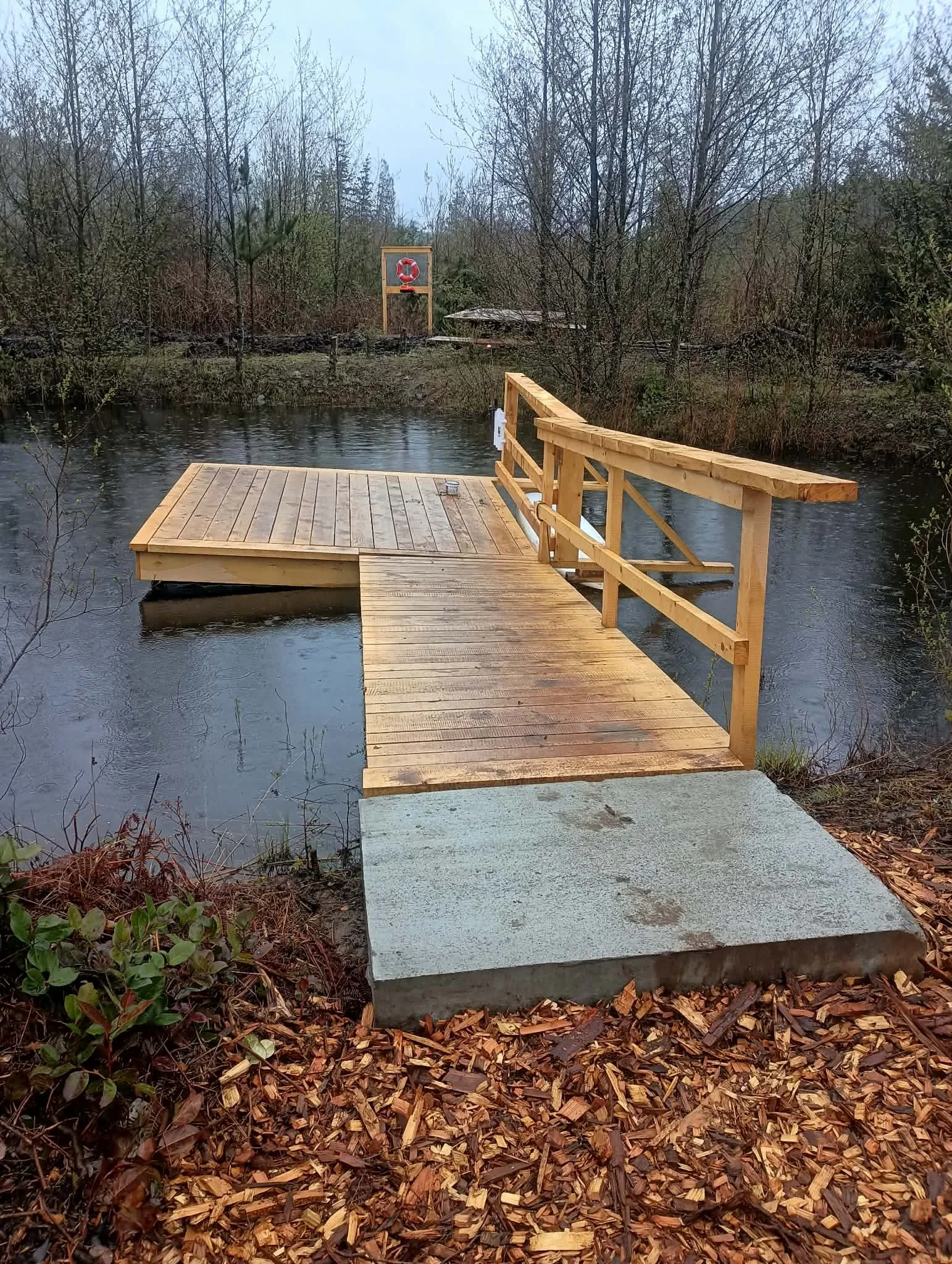 A wooden dock extends over a body of water surrounded by leafless trees and fallen leaves on the ground. A life preserver is mounted on a stand in the background.
