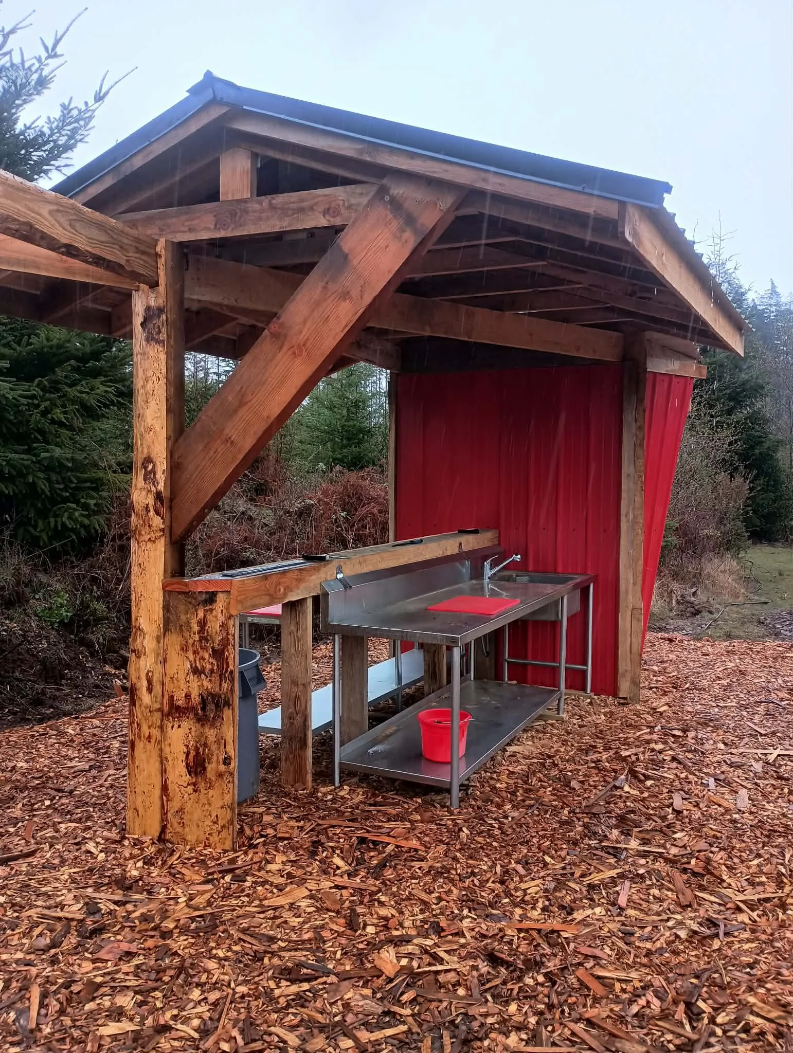 An outdoor wooden shelter with a metal sink and a red bucket underneath. The shelter has a roof and is located on a ground covered with wood chips, surrounded by trees and bushes.