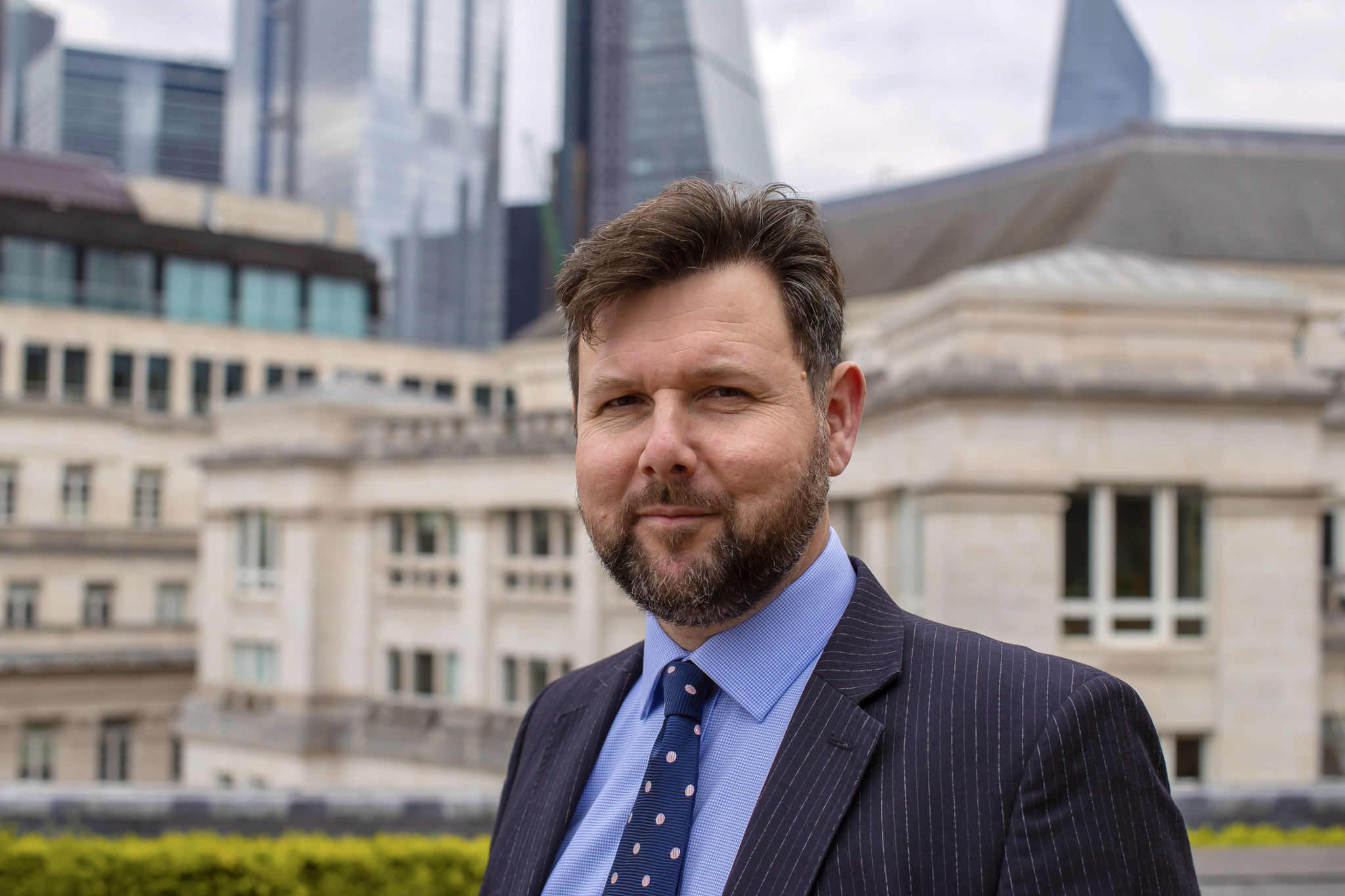 A man with a beard and brown hair in a suit with a blue shirt and polka dot tie standing outdoors in front of a cityscape with modern glass buildings and older stone structures.