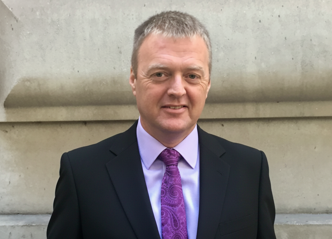 A man dressed in a black suit with a purple shirt and tie, standing outdoors in front of a stone wall.