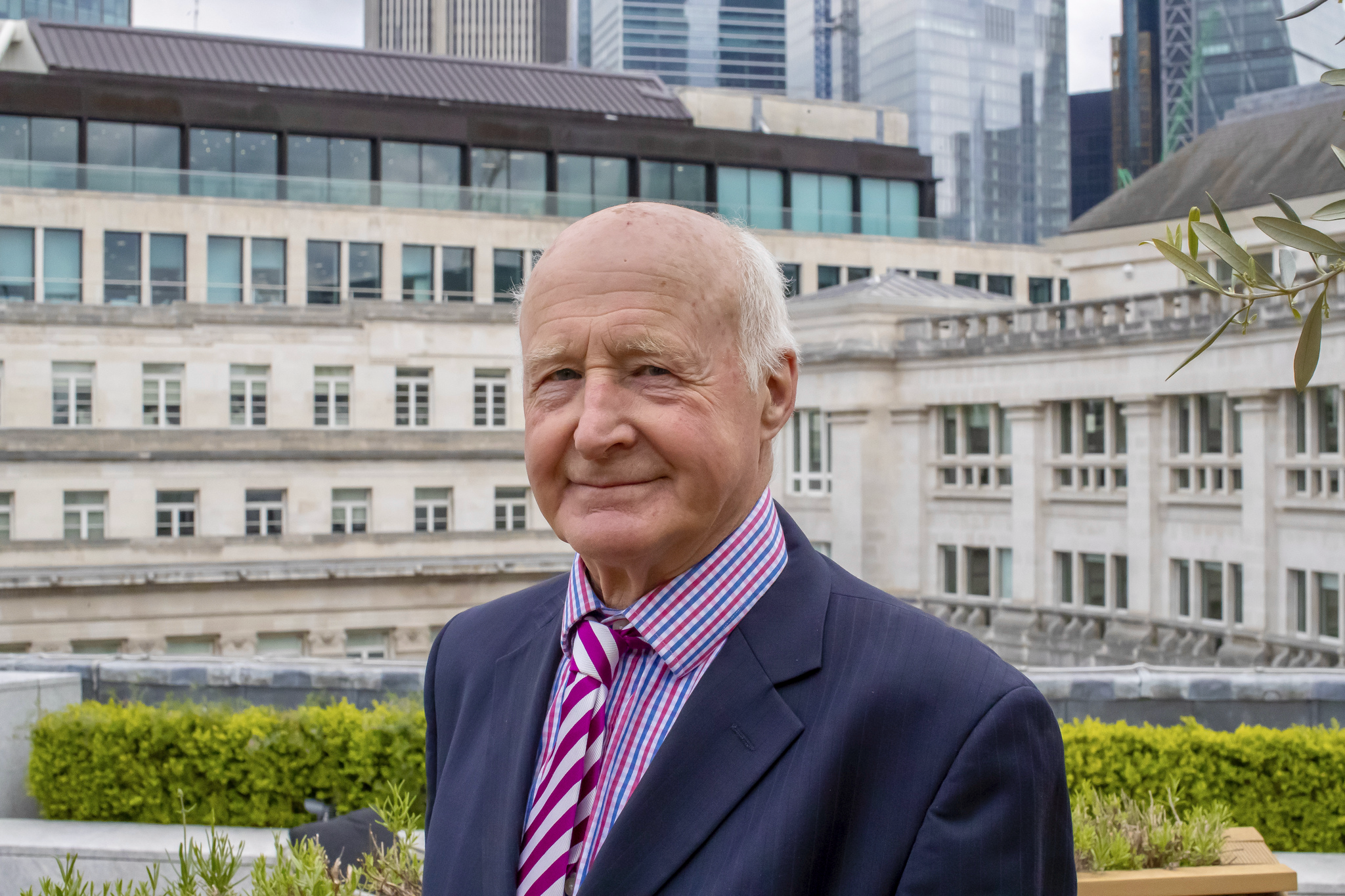 A man in a suit and striped shirt with a purple and white striped tie standing outdoors on a city rooftop or terrace with modern buildings, green plants, and a few trees in the background.