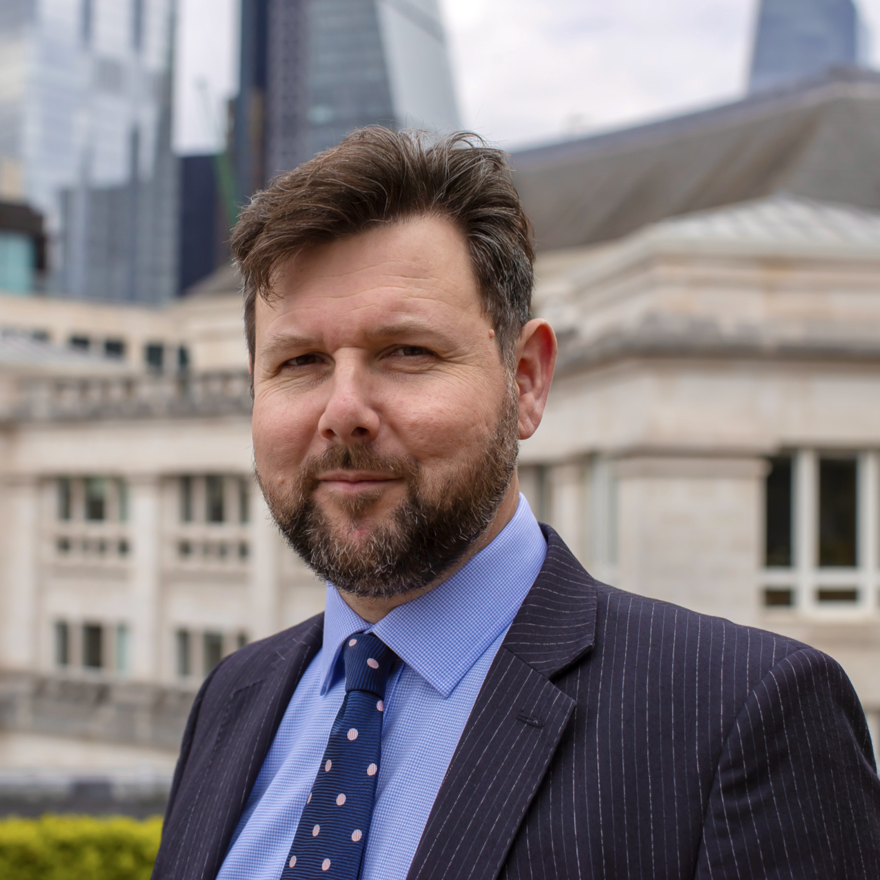 A man with a beard and brown hair in a suit with a blue shirt and polka dot tie standing outdoors in front of a cityscape with modern glass buildings and older stone structures.