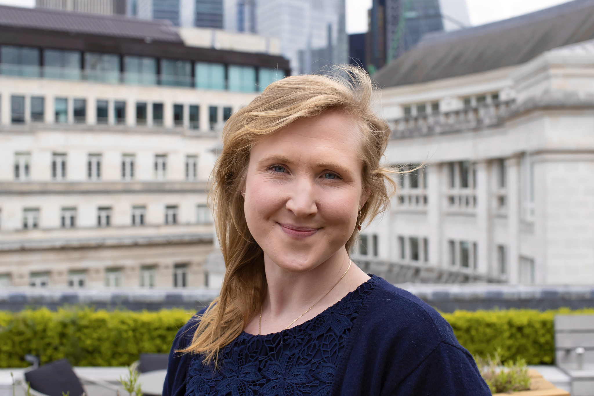 A woman with blonde hair and blue eyes is smiling outdoors with city buildings in the background.