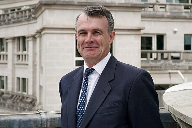 A man in a dark suit with a white shirt and patterned tie standing outdoors on a balcony with a stone building in the background.