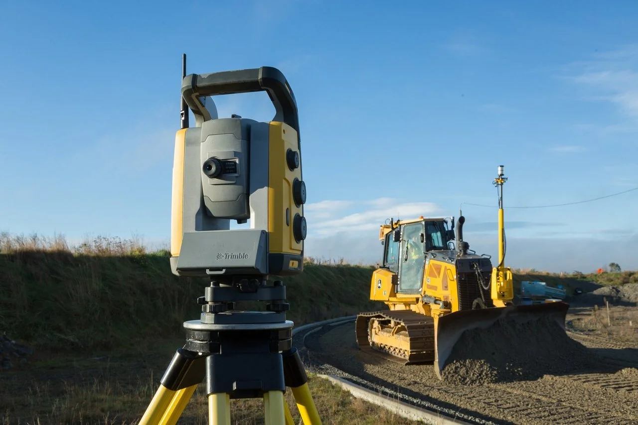 Construction site with a yellow bulldozer and a surveyor's tripod with a device on top, under a clear blue sky.
