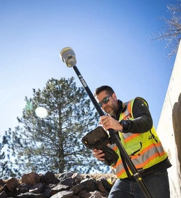 A man wearing sunglasses and a reflective safety vest is using surveying equipment outdoors under a clear blue sky with a large tree in the background.