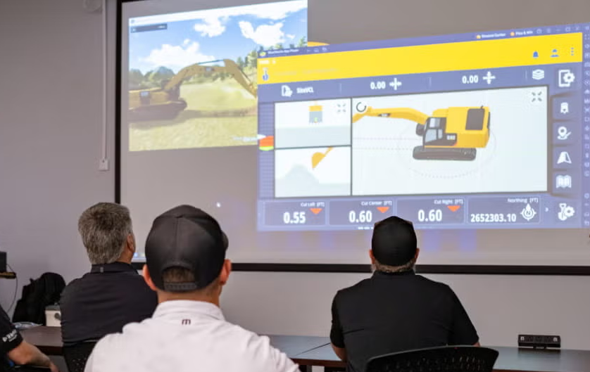 People watching a presentation on construction equipment operation simulation featuring a yellow excavator displayed on a screen.