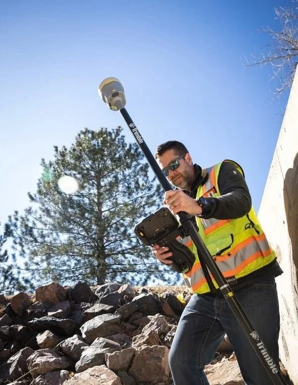A man wearing sunglasses and a high-visibility safety vest operates a drone controller with a long pole attached, outdoors in a rocky area under a clear blue sky.