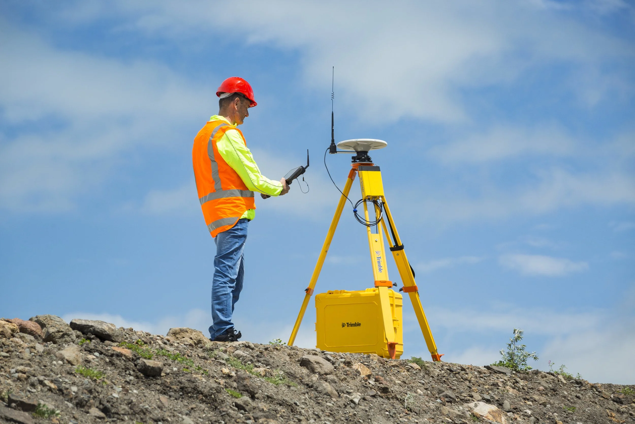 A surveyor in a red hardhat and high-visibility orange vest using a handheld device near a yellow surveying instrument on a tripod outdoors under a blue sky.