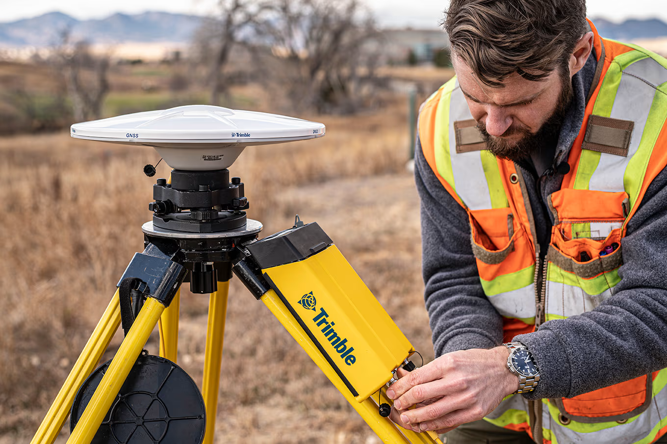 A surveyor setting up a GNSS surveying device in a field with dry grass and trees in the background.