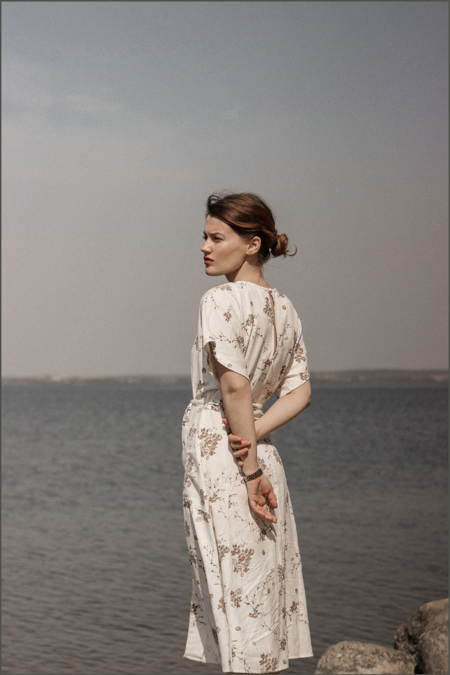 A woman with a bun hairstyle wearing a white floral dress standing by a body of water with a gray sky in the background.