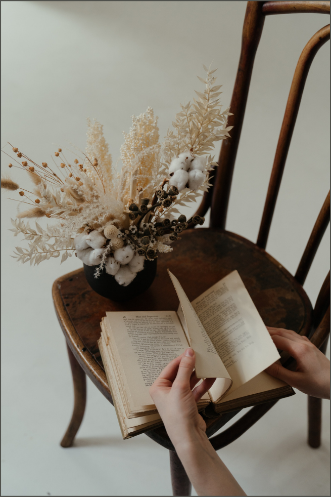A person's hands holding an open book models a vintage or classic style. The book is resting on an old wooden chair, and a black vase filled with dried flowers, including cotton stems, is positioned on the chair's seat. The setting has a minimalist and rustic aesthetic.