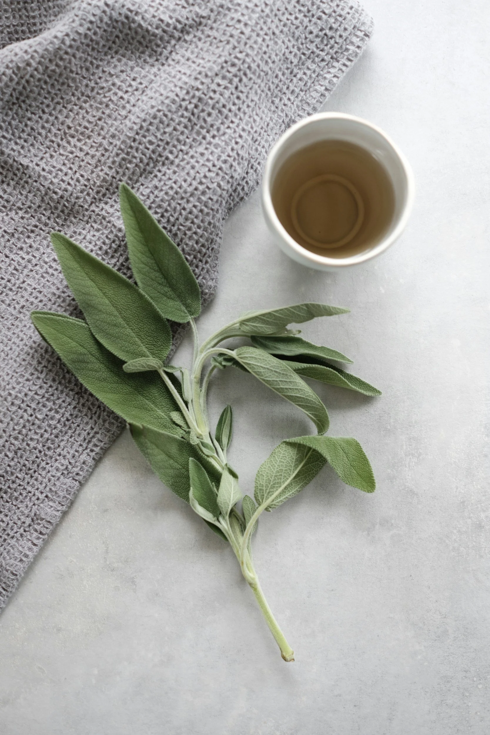 Fresh sage leaves with a cup and a textured cloth on a light surface.