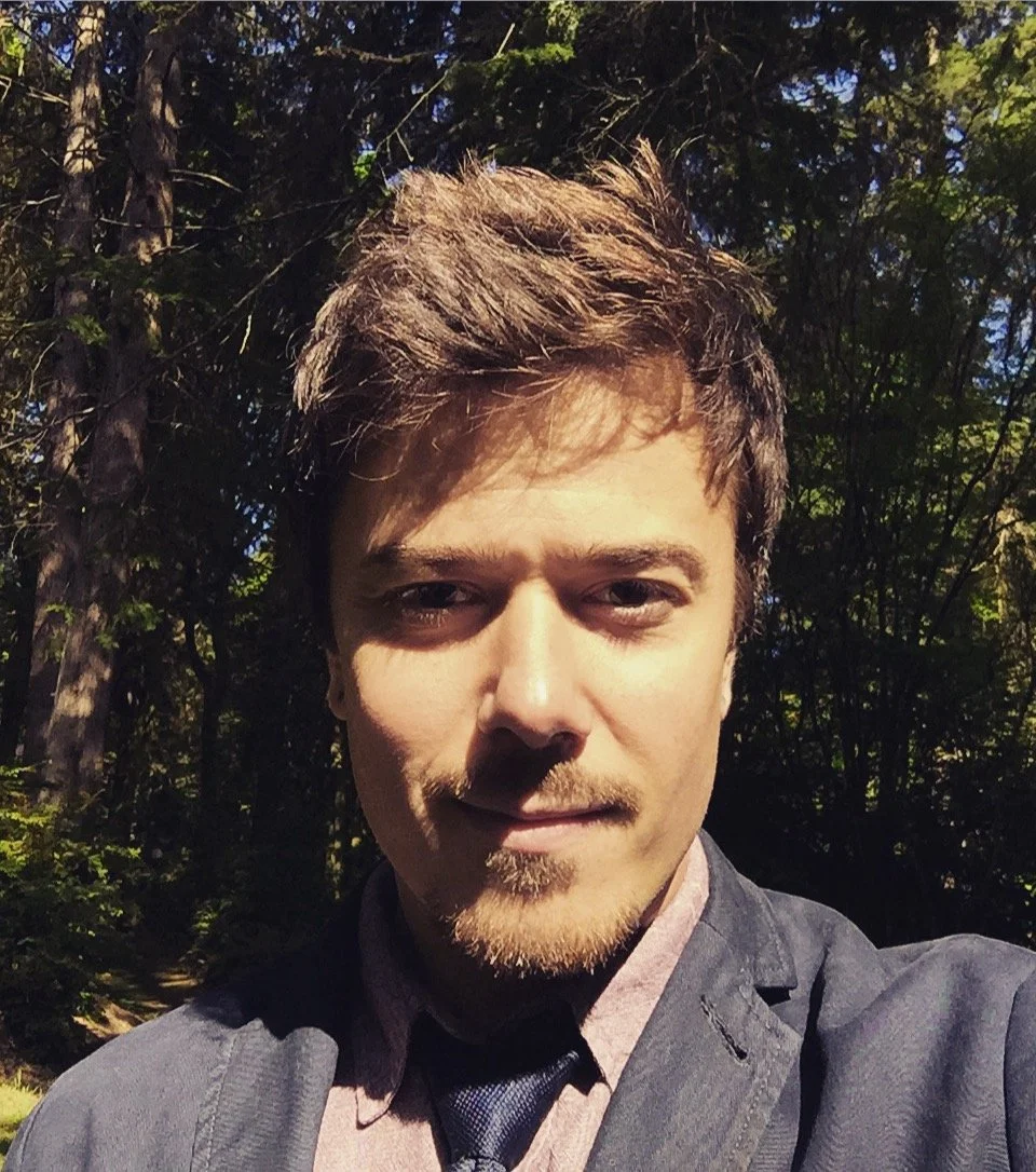 A young man with light brown hair, a goatee, and light skin taking a selfie outdoors in a wooded area, with trees and sunlight in the background.
