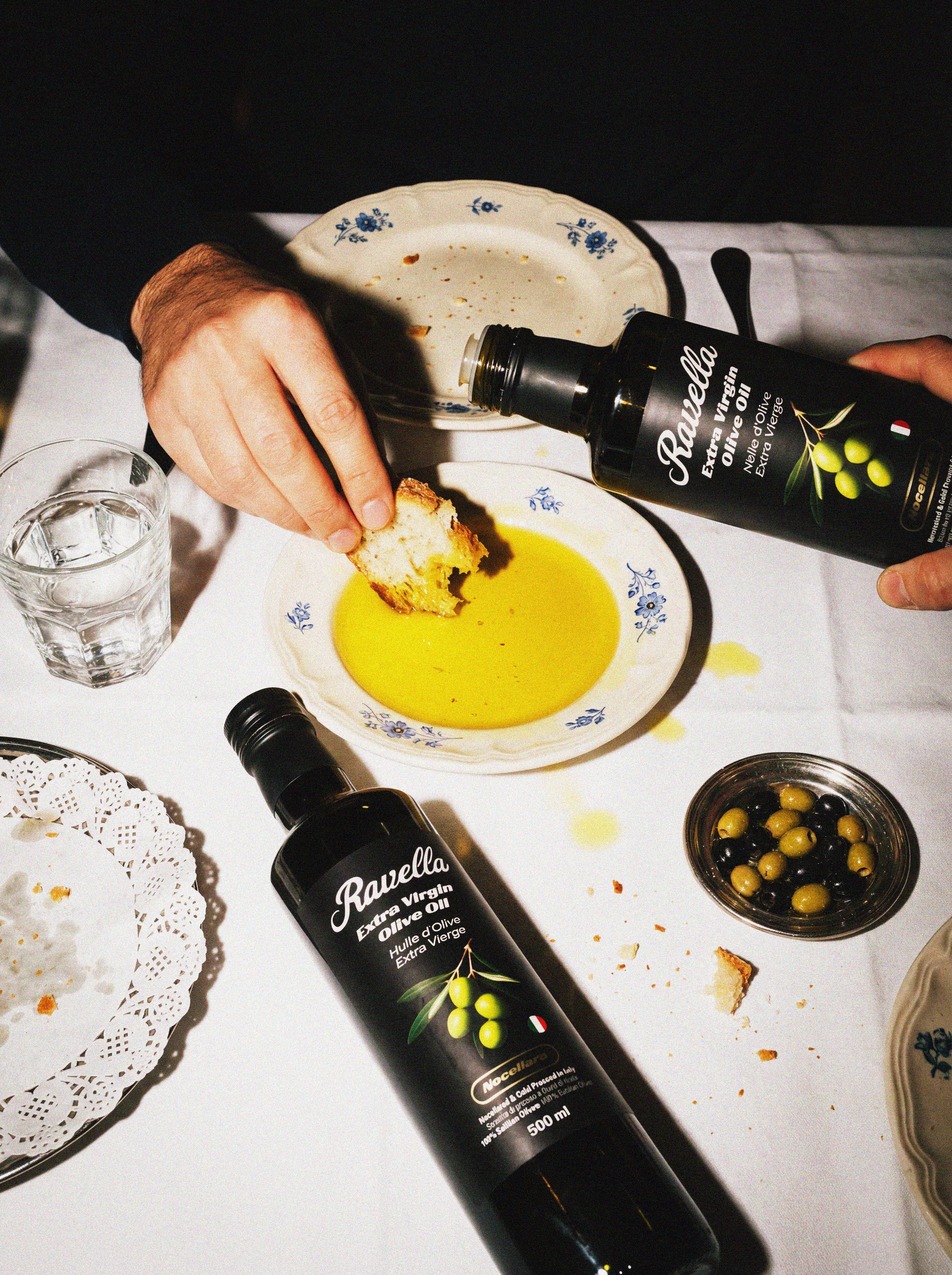 A person pours extra virgin olive oil from a  ravella bottle onto a piece of bread dipped in olive oil on a white plate. The table has a glass of water, a small bowl of green and black olives, and additional plates, with a white tablecloth.