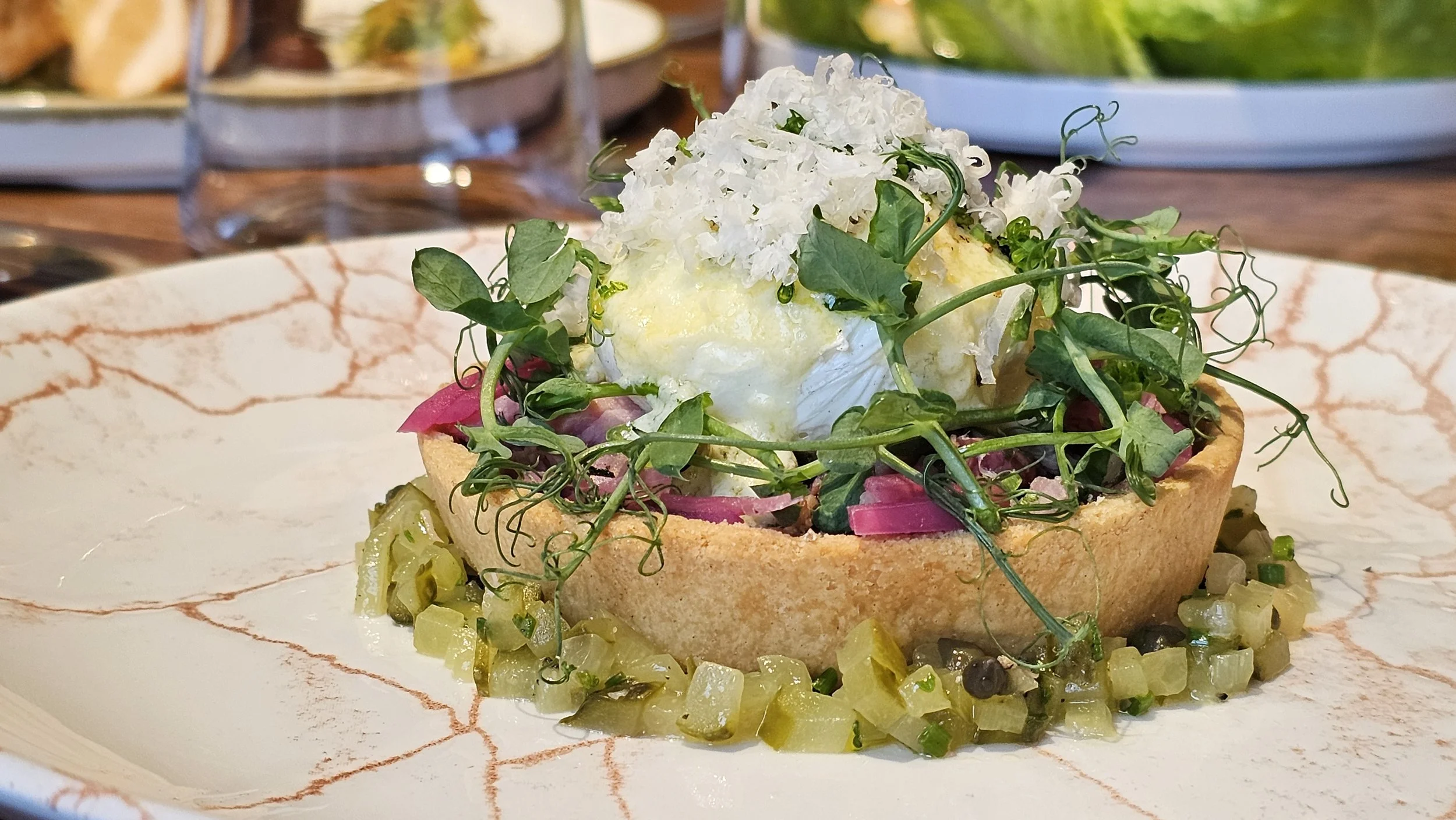 A savory tart topped with greens, edible flowers, and a poached egg, surrounded by diced pickles and herbs on a decorative plate.