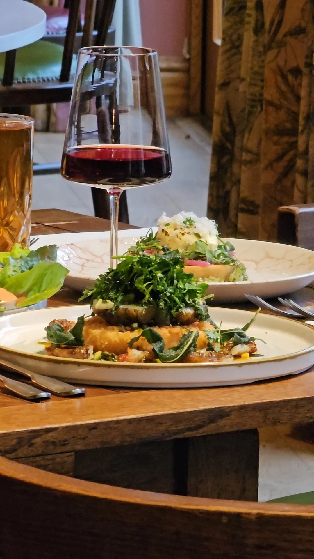 A glass of red wine, a plate with food garnished with greens, and another dish with mashed potatoes and vegetables on a wooden table in a restaurant.