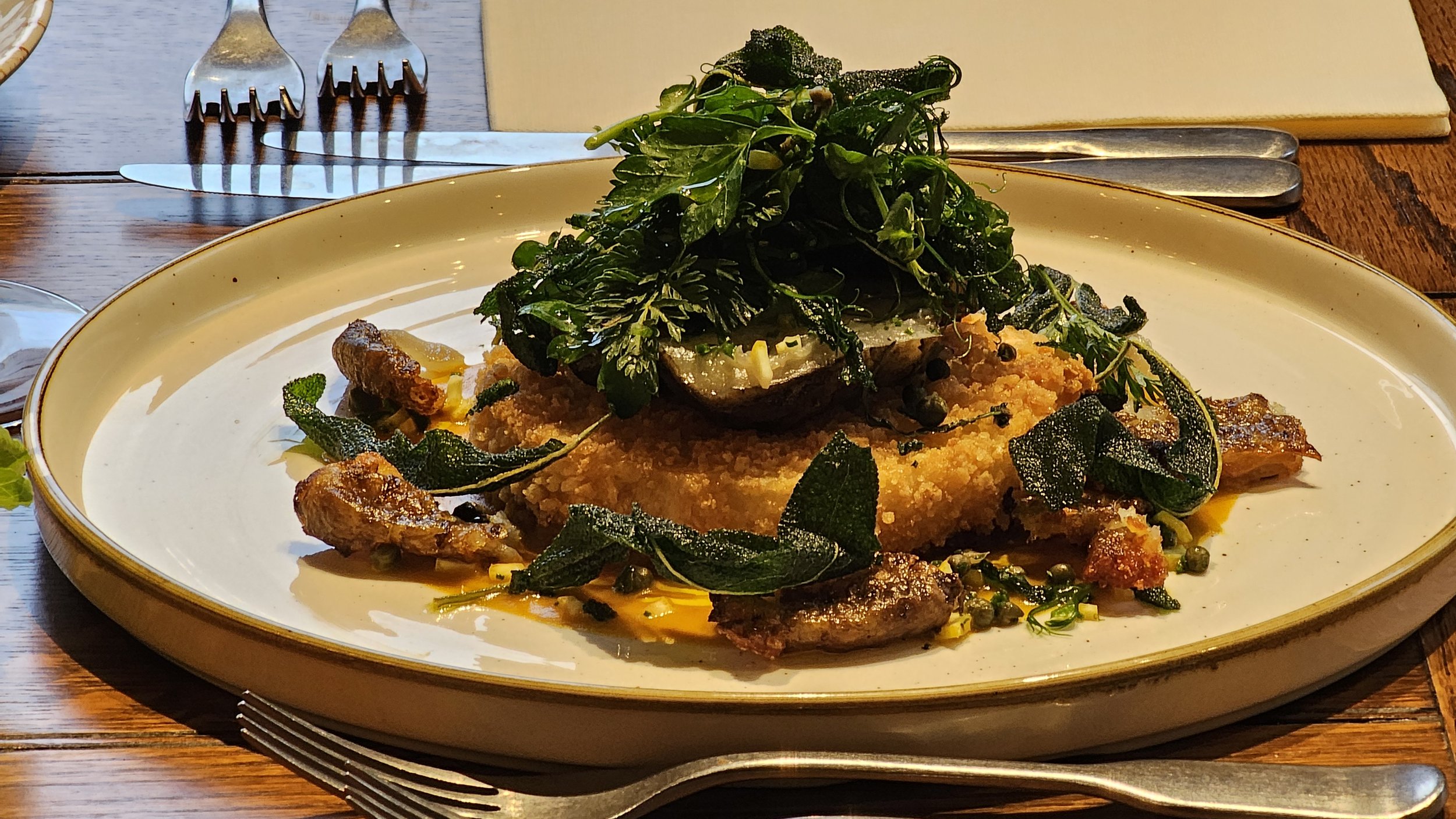 A plated dish featuring fried fish fillet topped with fresh greens, lime wedge, and dark leafy herbs, served on a white oval plate.