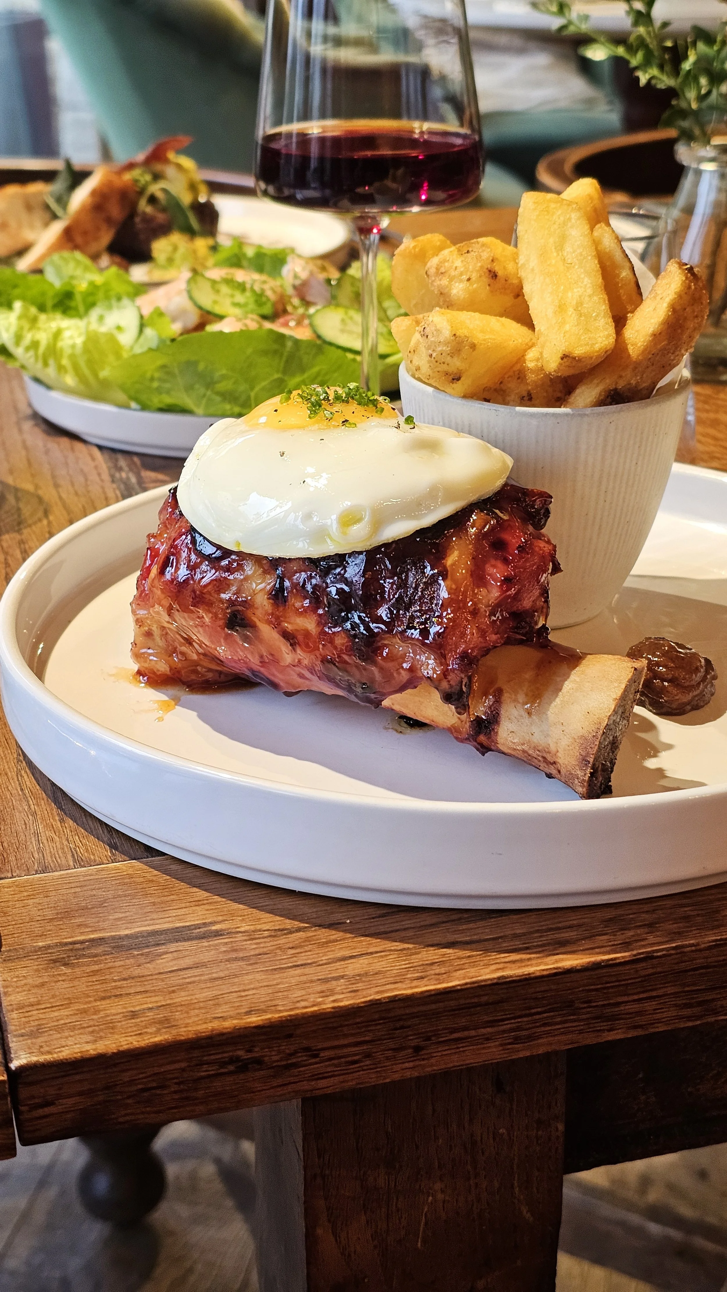 A plate with a serving of roasted pork with a fried egg on top, a side of thick-cut fries, and a small bowl of salad in the background with a glass of red wine.