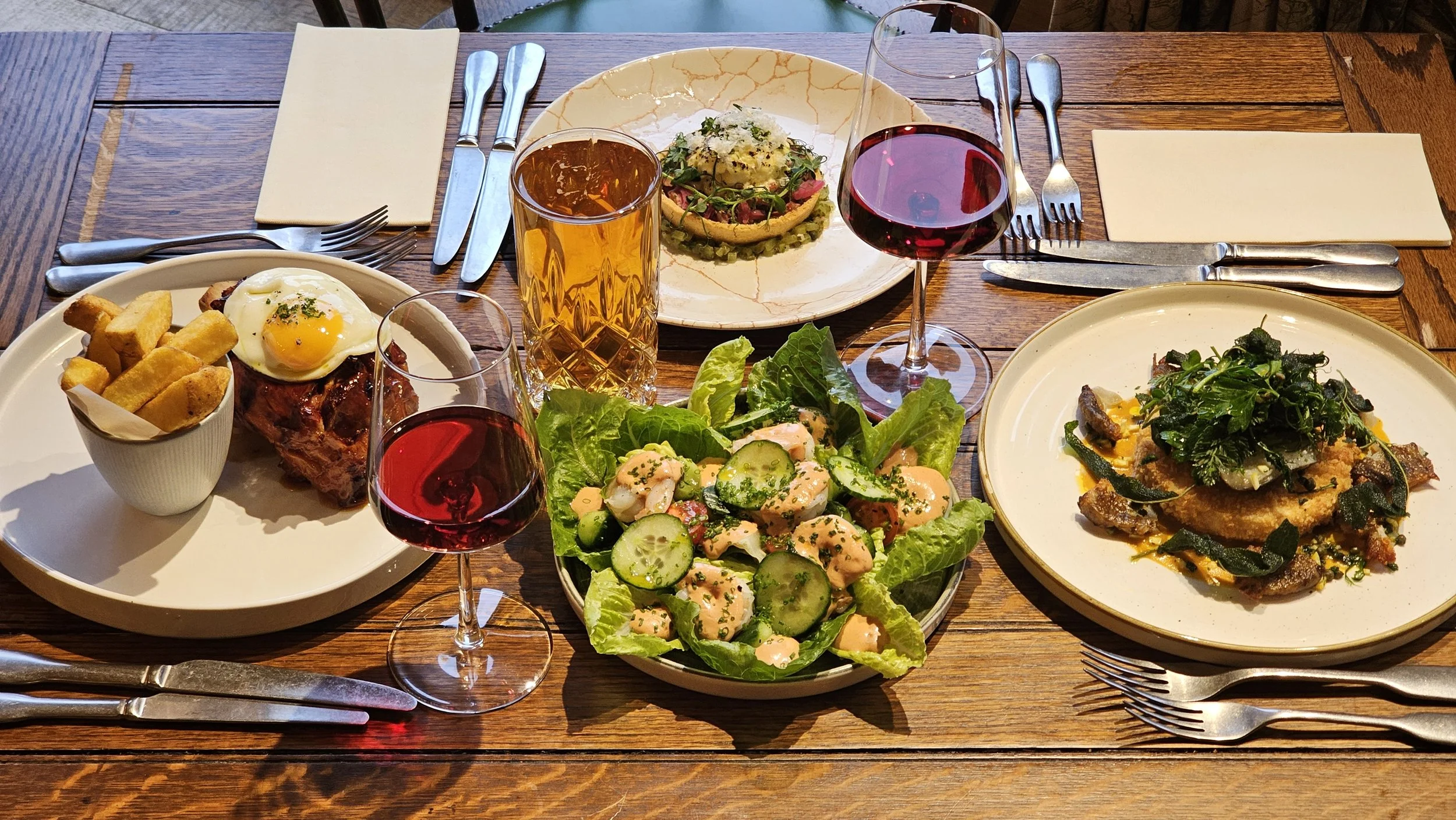 A wooden dining table set with various dishes including a salad, steak with a fried egg and fries, fish with greens, and a small appetizer plate with a colorful garnish. There are two glasses of red wine, one with a large bowl and the other with a sm