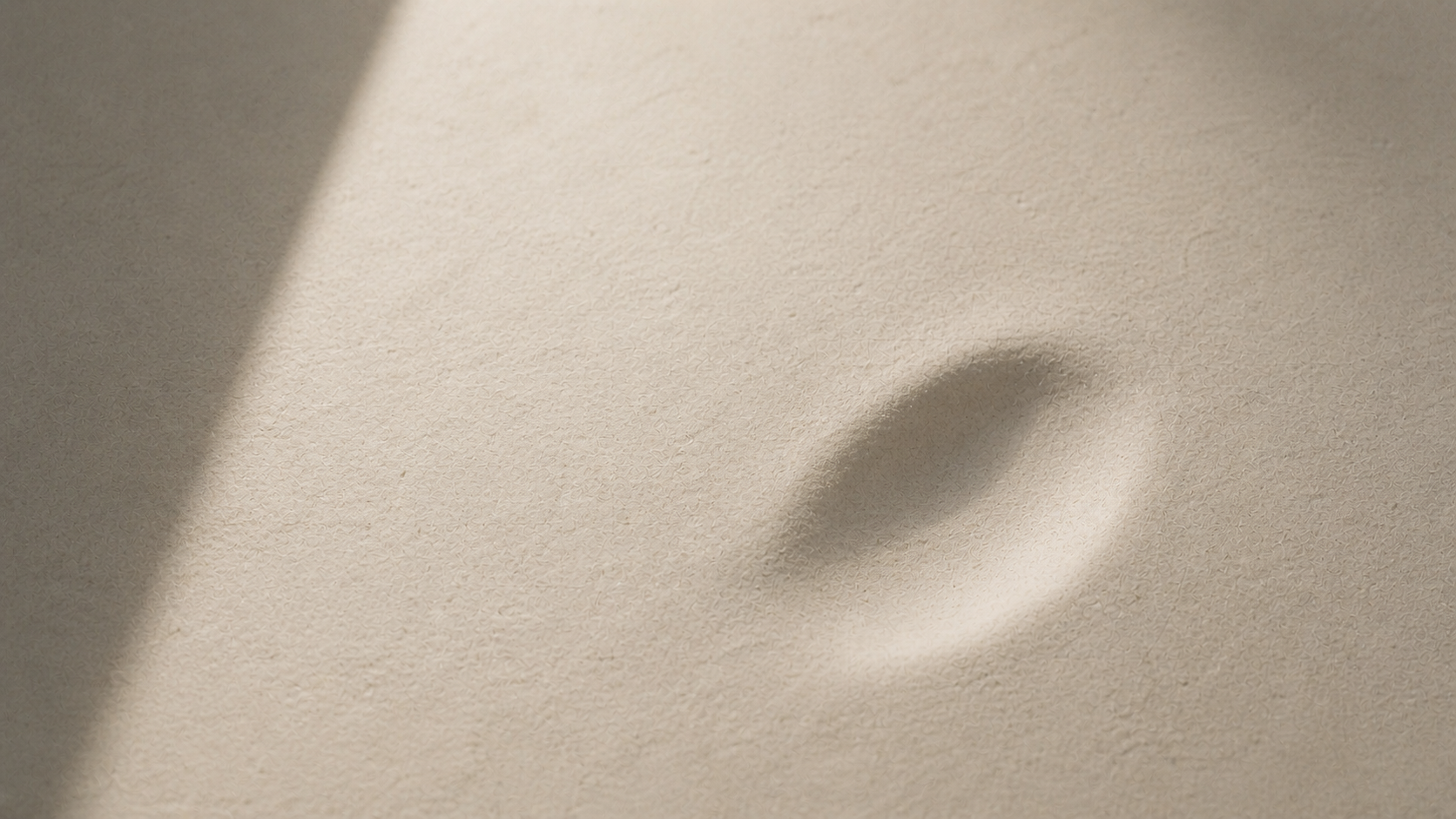 Close-up of a textured pale surface with a soft indentation, lit from one side to reveal subtle shadow and depth.