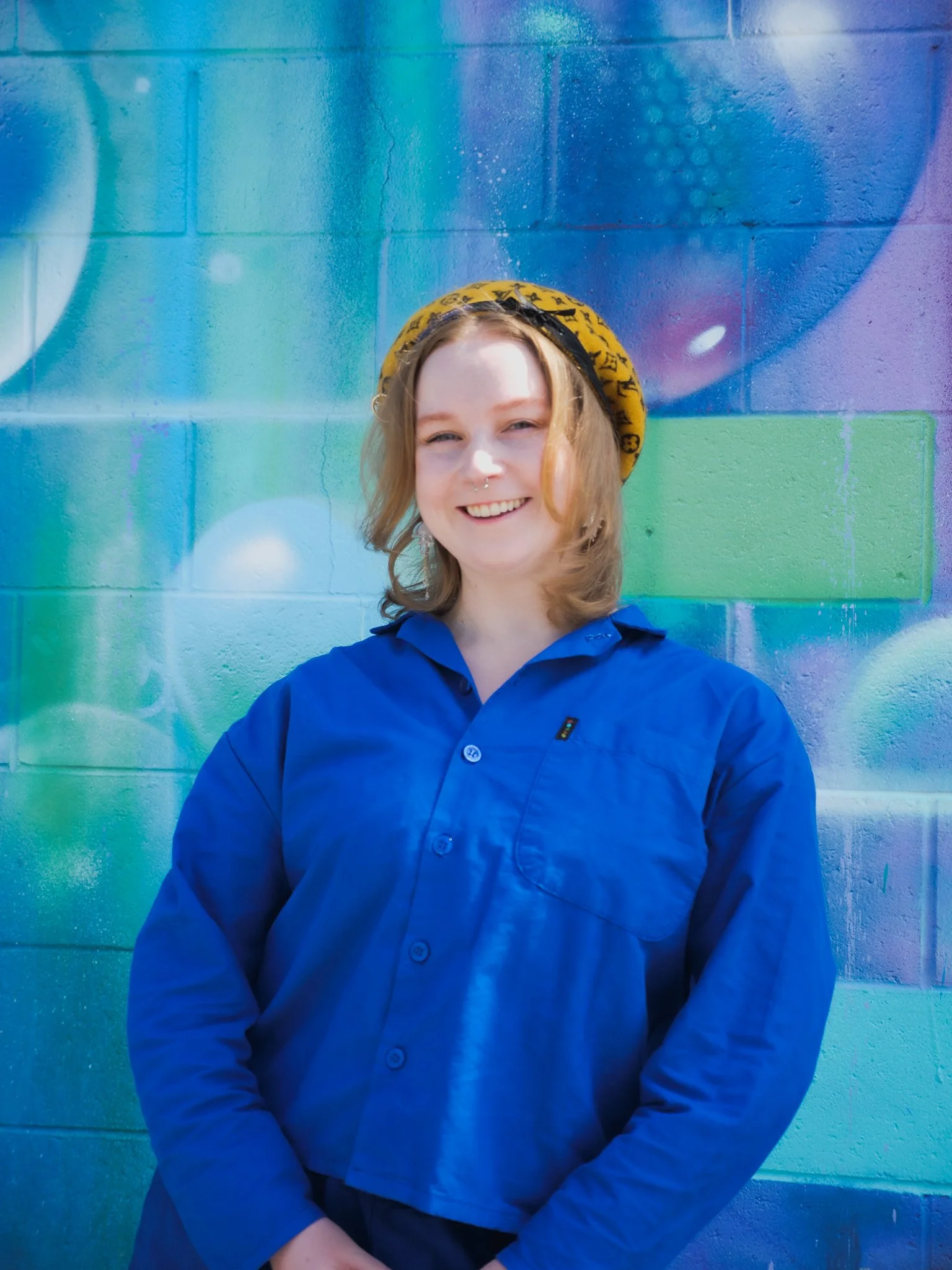 A young woman with shoulder-length hair smiling, wearing a yellow patterned beret, a blue button-up shirt, and silver earrings, standing in front of a colorful mural on a brick wall.