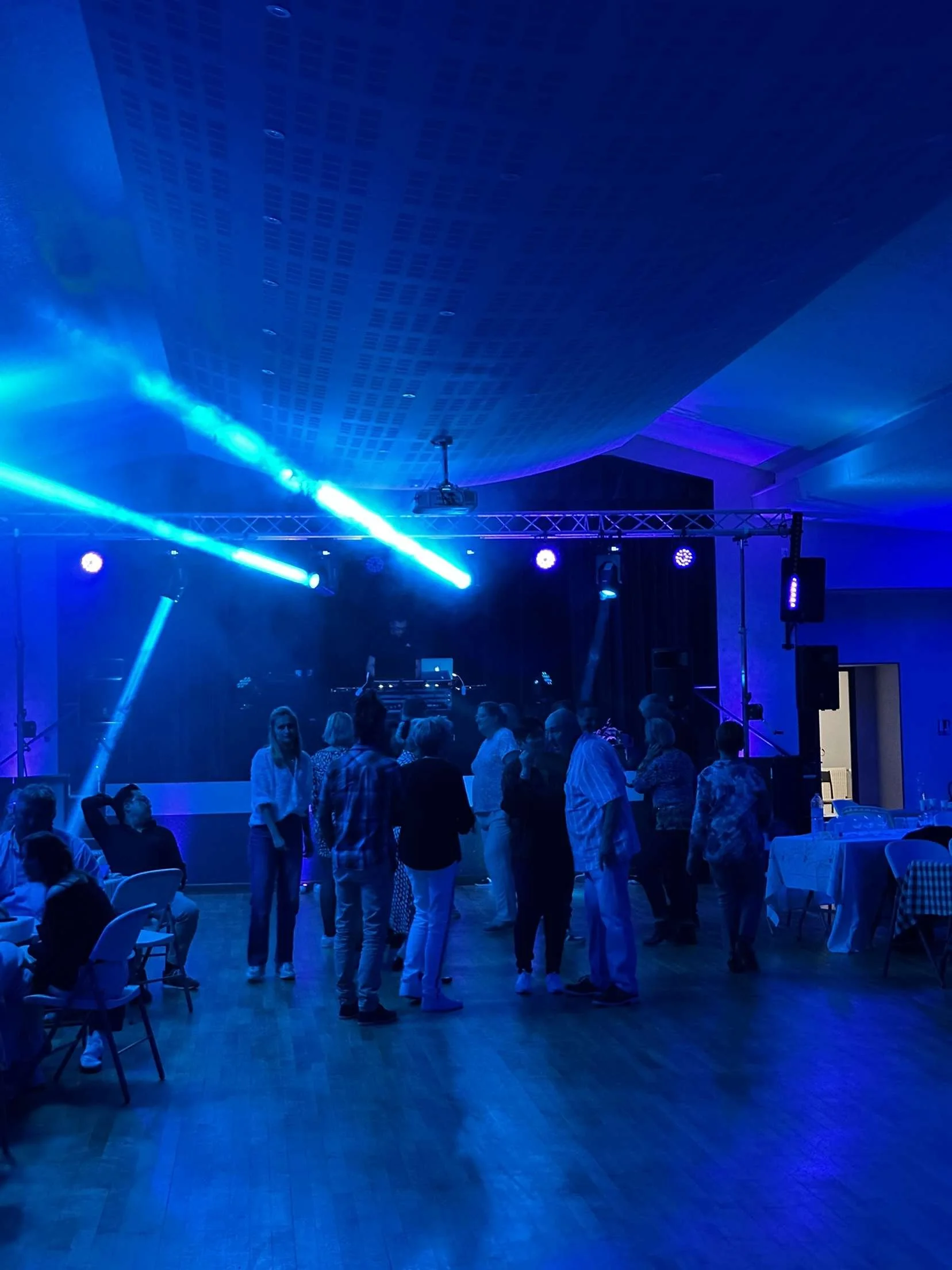 Groupe de personnes dans une salle de fête avec éclairage bleu, dansant et socialisant devant une scène avec équipements audio et éclairages LED.
