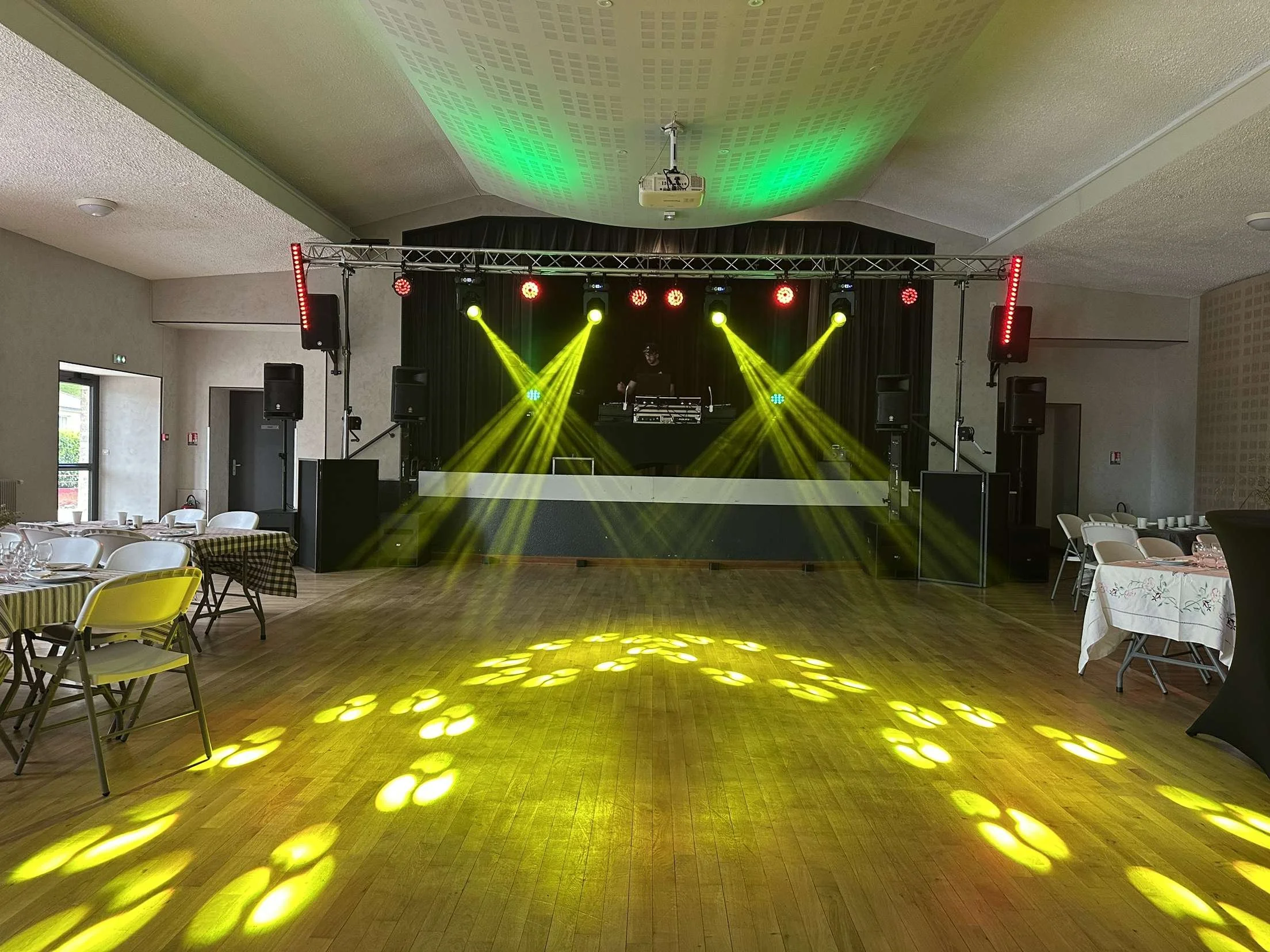 Salle de fête ou événement avec un espace vide au centre pour danser, un DJ sur scène avec un éclairage de scène jaune et rouge, des tables et chaises autour avec de la vaisselle, et un éclairage de piste de danse jaune projeté au sol.