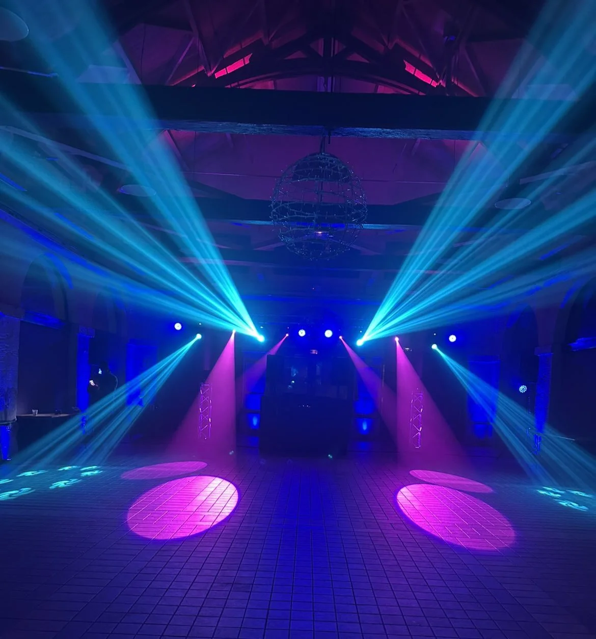 Salle de danse ou de concert avec éclairage coloré, projecteurs bleus et roses, sphère suspendue au plafond, sol en carreaux, ambiance festive.
