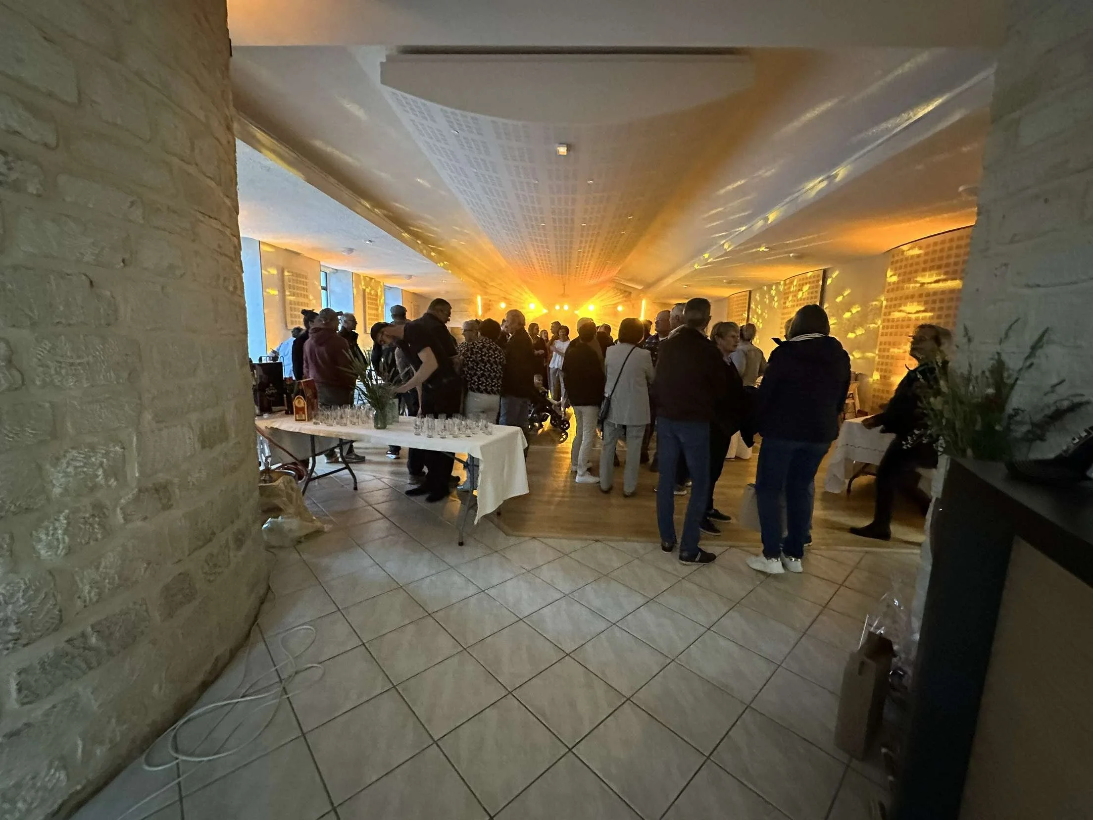 Une salle de réception avec des personnes discutant, un buffet avec des verres, et un décor chaleureux avec des lumières dorées.