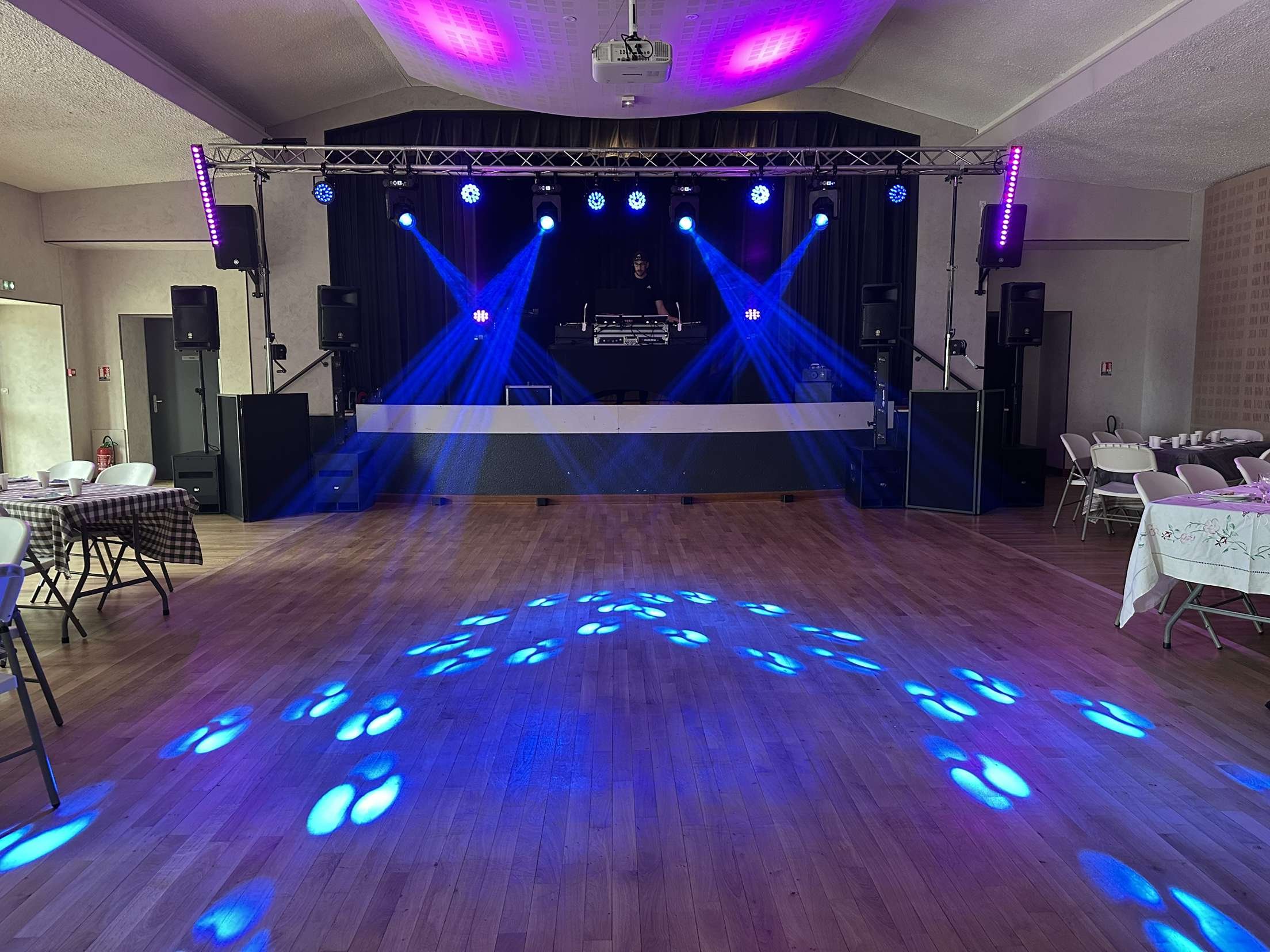 Salle de fête avec une scène équipée de lumière bleue et de haut-parleurs, tables et chaises sont disposées autour avec des nappes à carreaux et des tables à fleurs