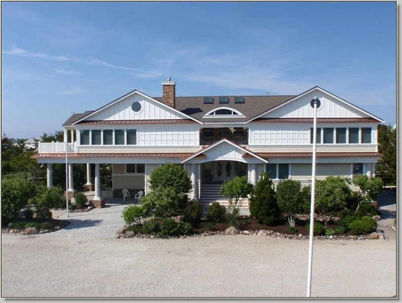 Large two-story house with a white exterior, multiple windows, and a front porch, surrounded by landscaping with small bushes and trees.