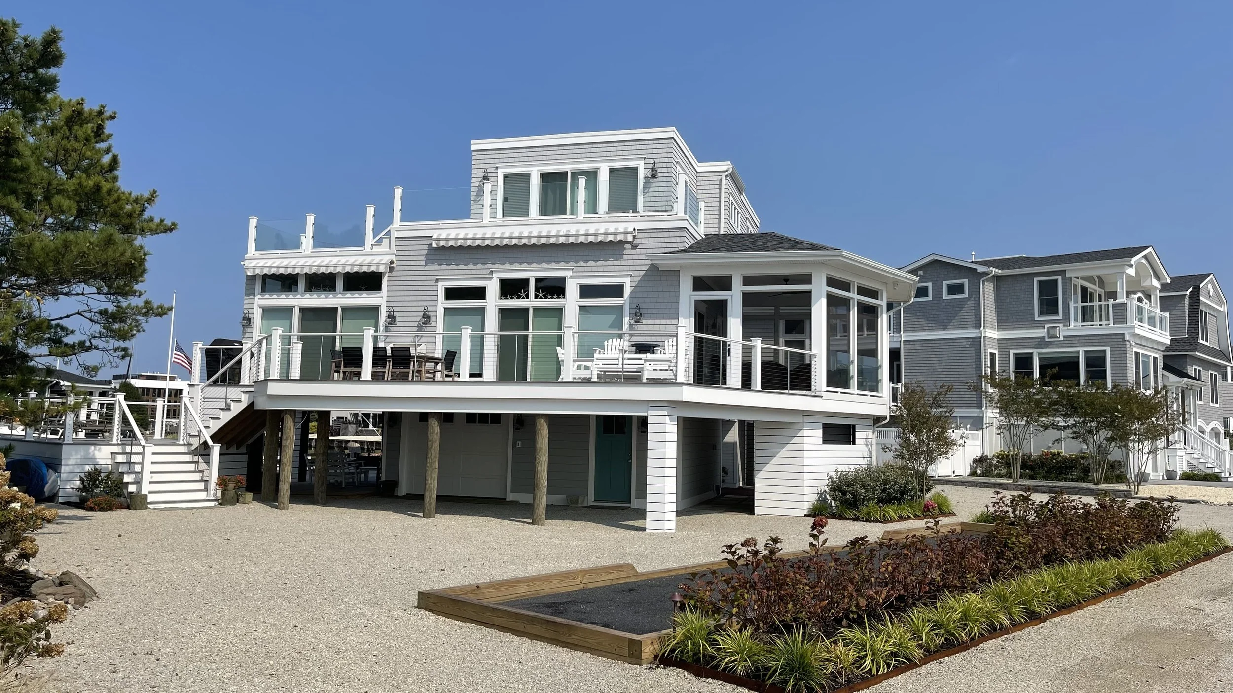 A multi-story beach house with a large deck, white siding, and glass railings, overlooking a gravel yard with landscaped garden beds.