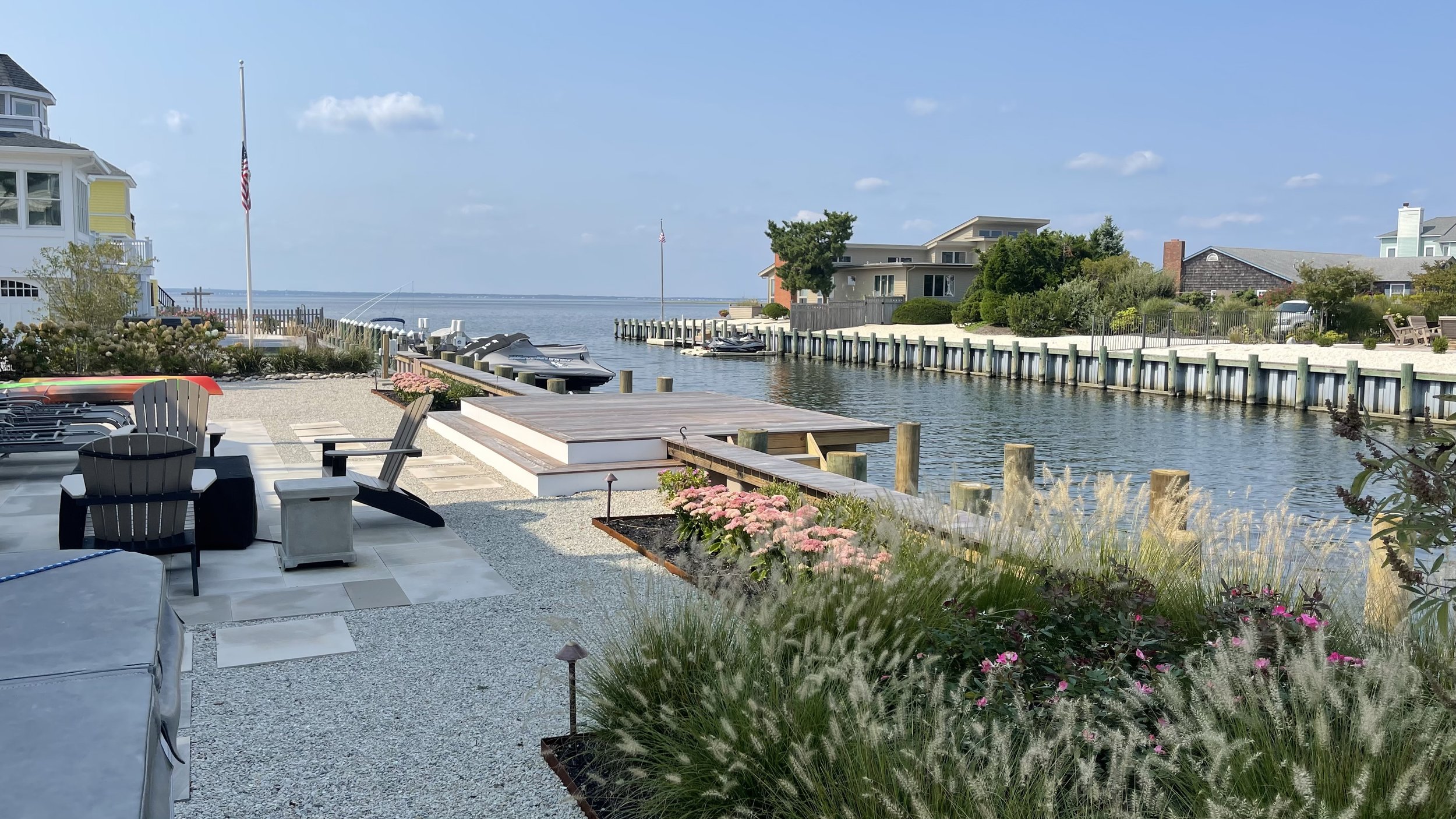 A backyard view by the water with a patio, Adirondack chairs, garden beds with pink flowers, boats docked at a pier, and houses in the background under a blue sky.