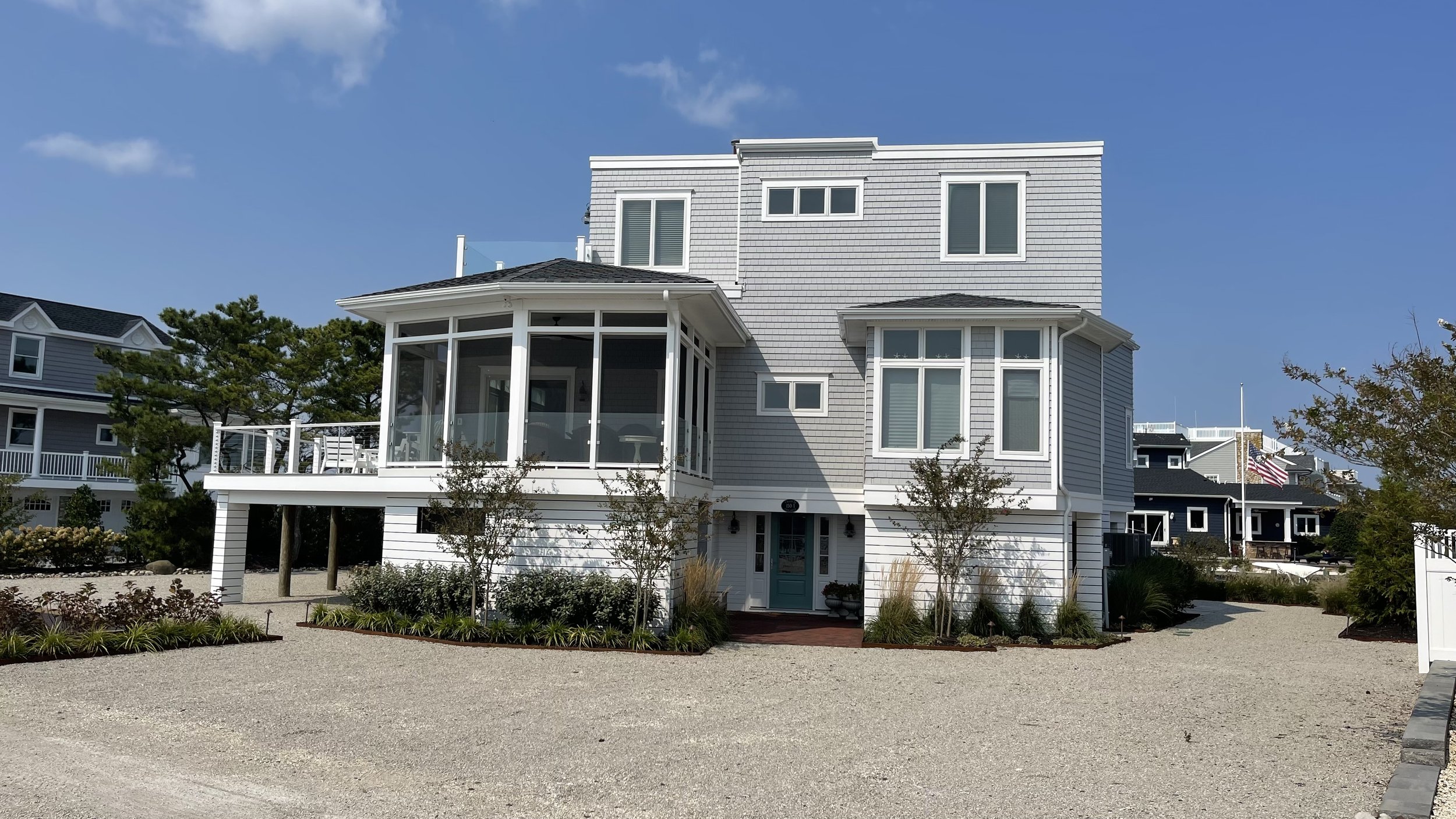 A large white multi-story beach house with a sunroom and bay windows, surrounded by landscaped trees and plants on a gravel pathway, under a bright blue sky.