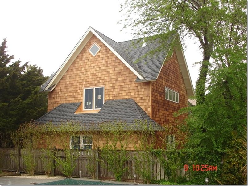 two-story house with brown shingle siding, gray gable roof, and multiple windows, surrounded by trees and a weathered wooden fence