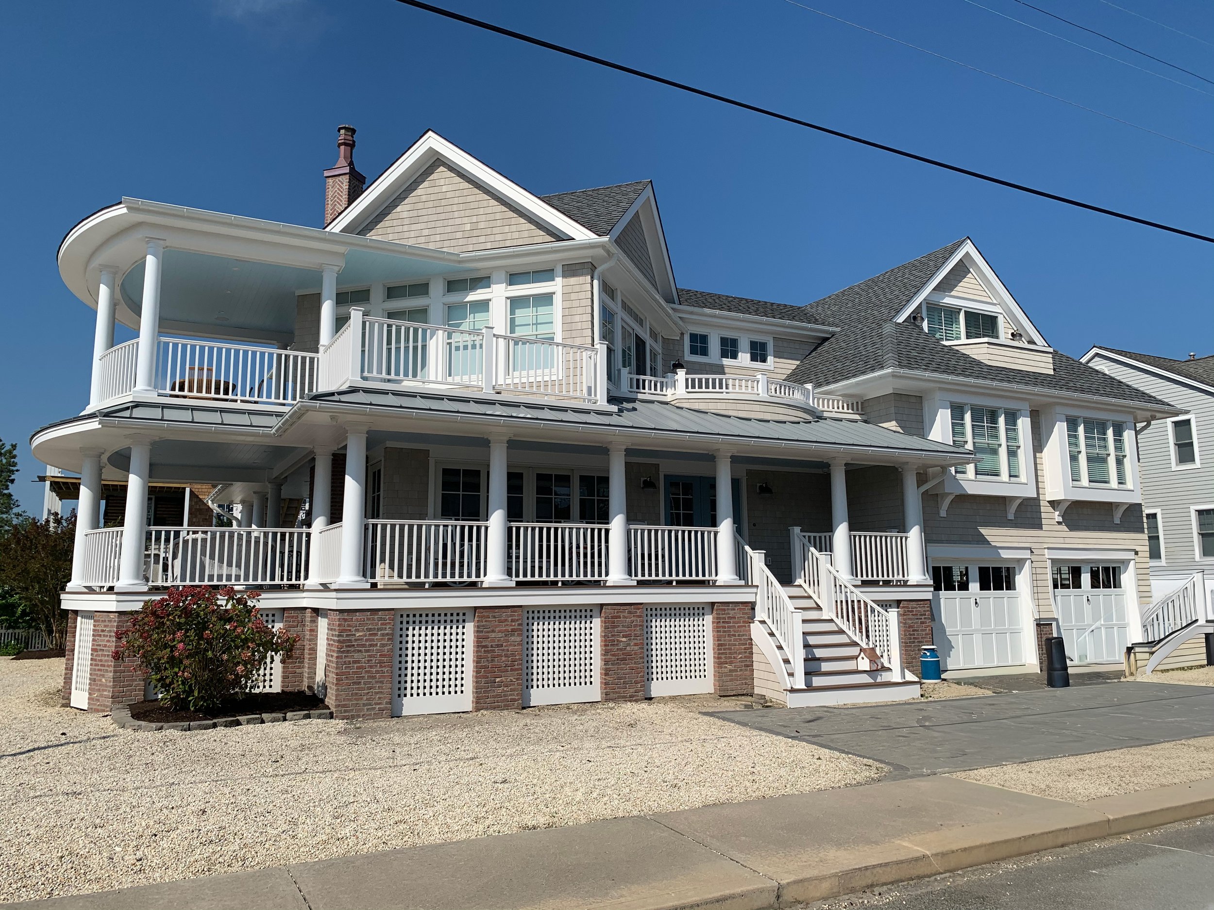 Large multi-story house with multiple balconies, white railings, and a brick foundation, set against a clear blue sky.
