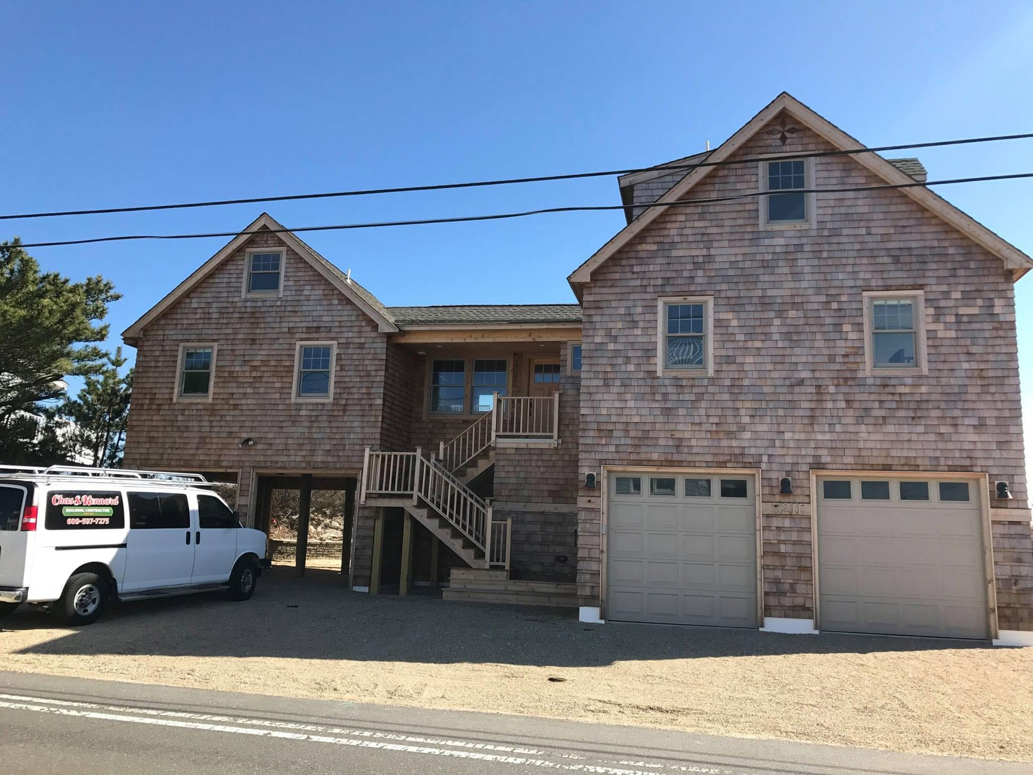 A large two-story house with wooden shingle siding, three garage doors, and a staircase leading to the front door. A white work van is parked in front.