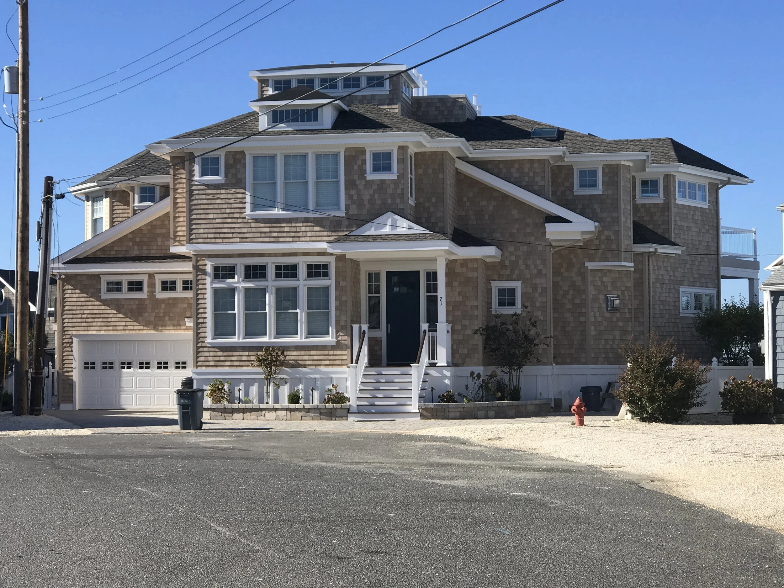 Large multi-story house with beige shingles, white trim, multiple windows, and a white garage door. The house has stairs leading to the front door and a small landscaped garden with bushes and trees in front. Power lines and a fire hydrant are visibl