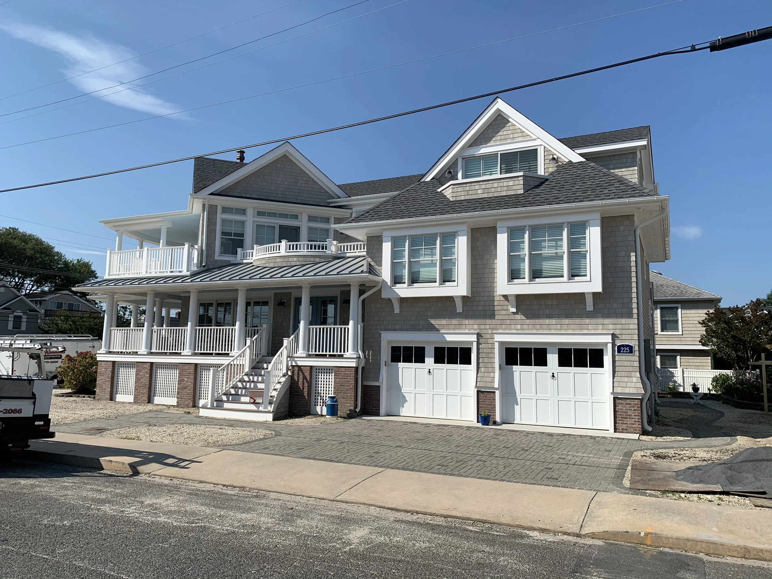 A large multi-story house with a beige shingle exterior, white trim, and a gray roof. The house has two attached garages with white doors, a front porch with stairs, and multiple windows. The sky is clear and blue.