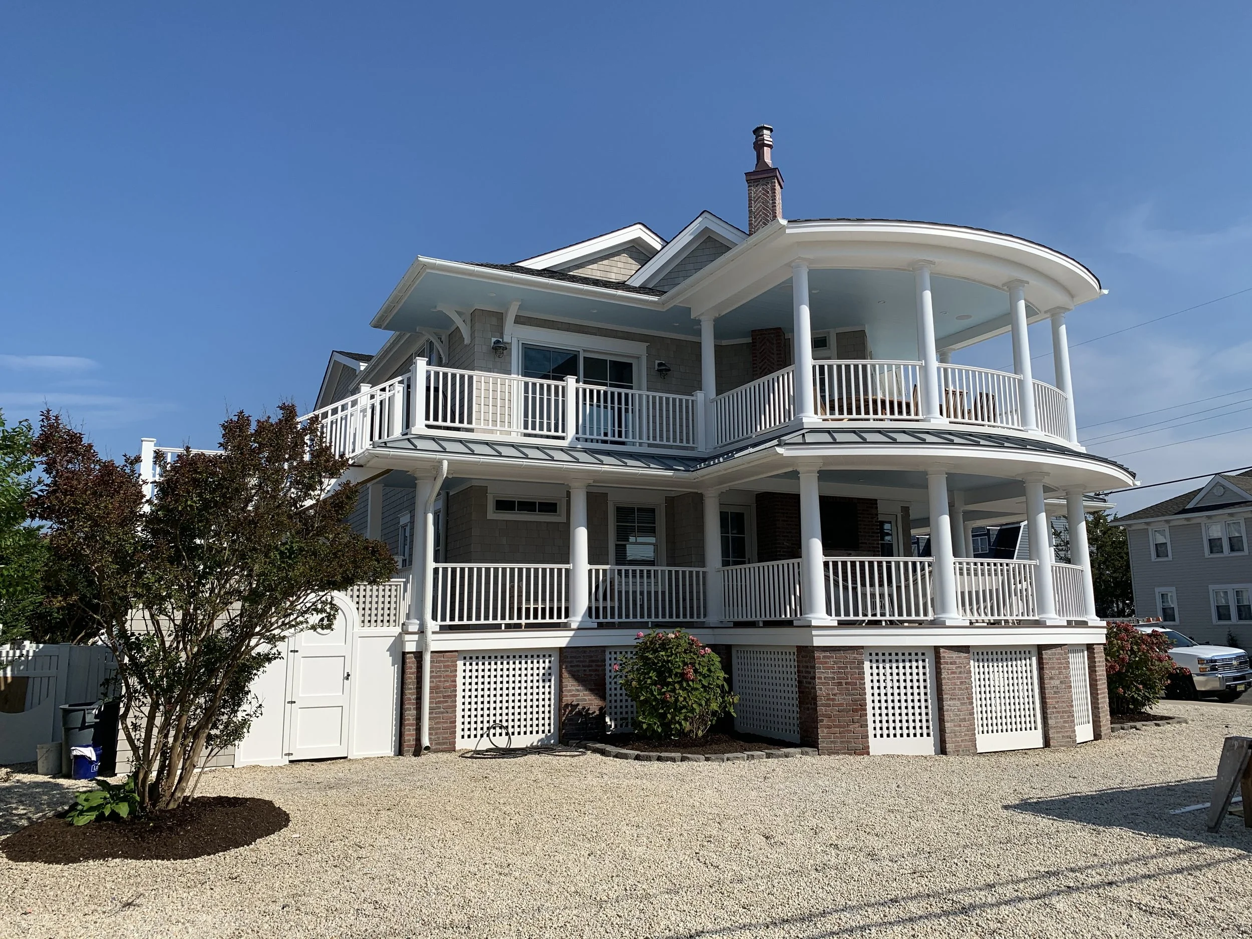 Multi-story house with round balconies, white railings, brick accents, and a gravel yard under clear blue sky.