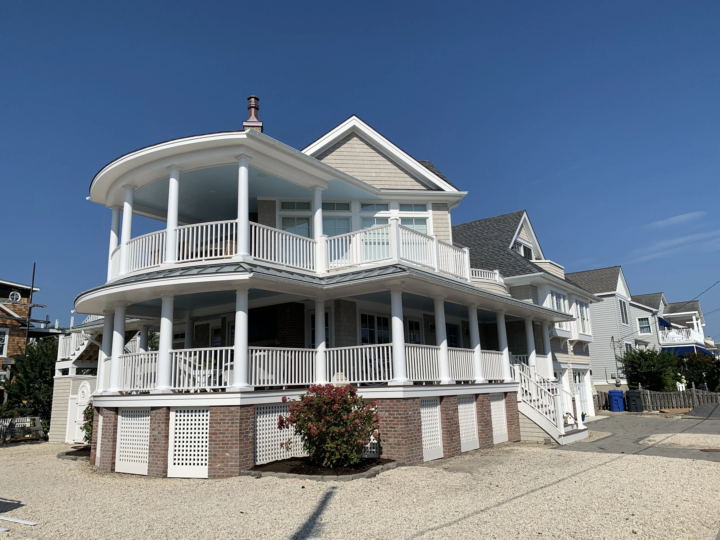 A large, multi-story house with white balconies and pillars, gray roof, brick foundation, and surrounding gravel yard under a clear blue sky.