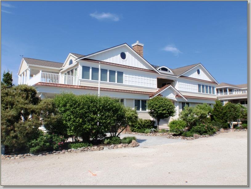 Large multi-story white house with multiple gable roofs, surrounded by green bushes and trees, under a clear blue sky.